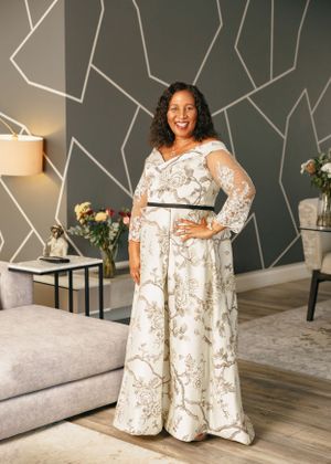 A full-length view of someone in a white floral dress with sheer sleeves against a geometric wall backdrop.