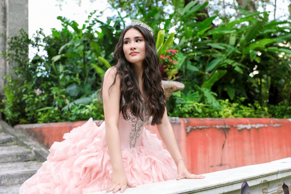 Model in coral pink gown poses elegantly on outdoor stone bench.