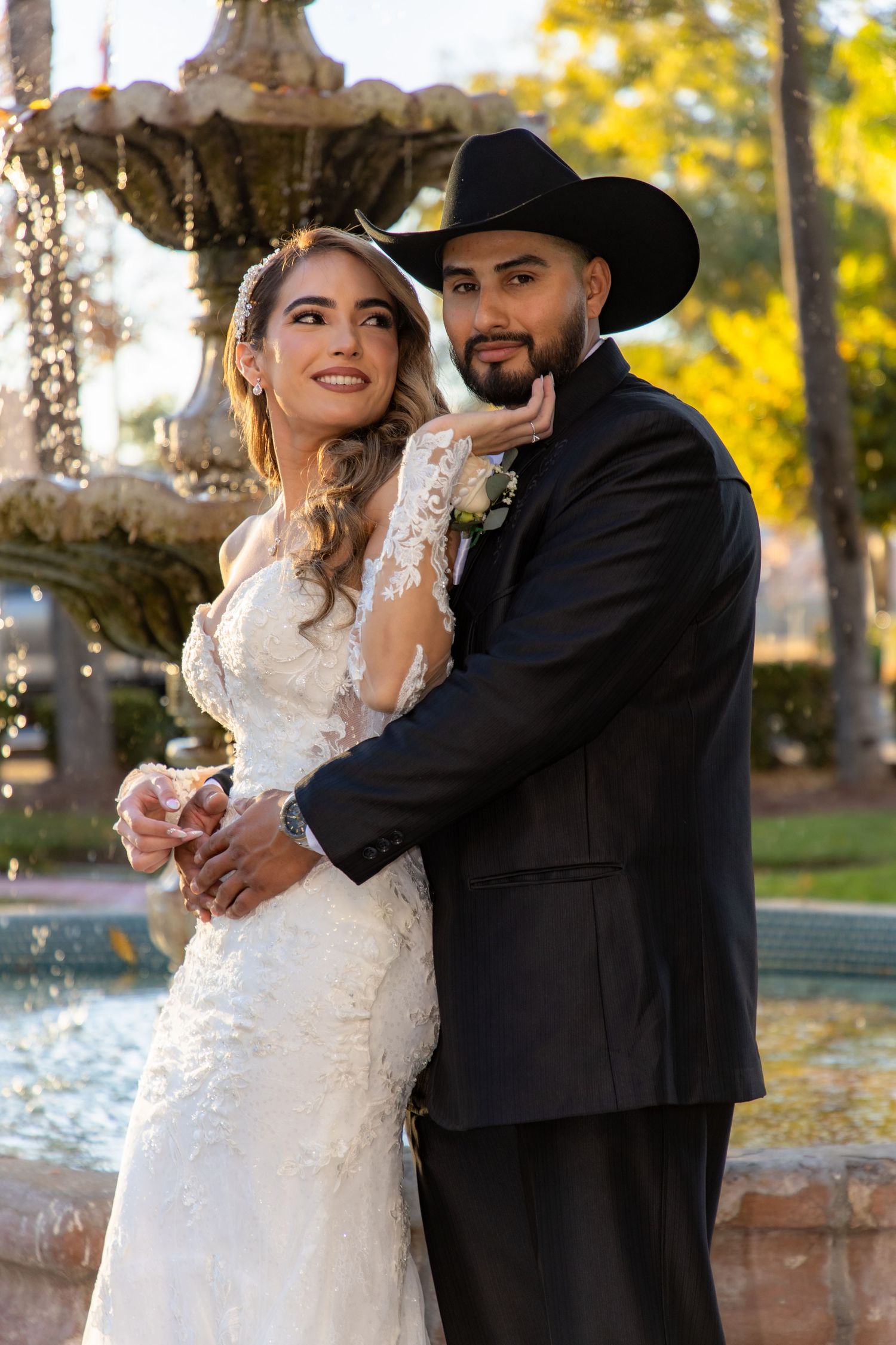 Elegant bride and groom in a black cowboy hat pose together near a fountain after their wedding.
