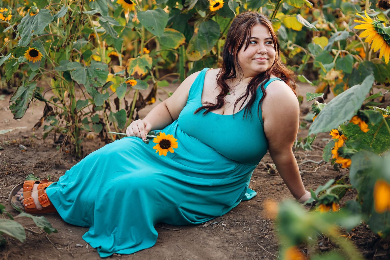 Woman in turquoise dress poses among blooming sunflowers in a garden setting. Senior photography
