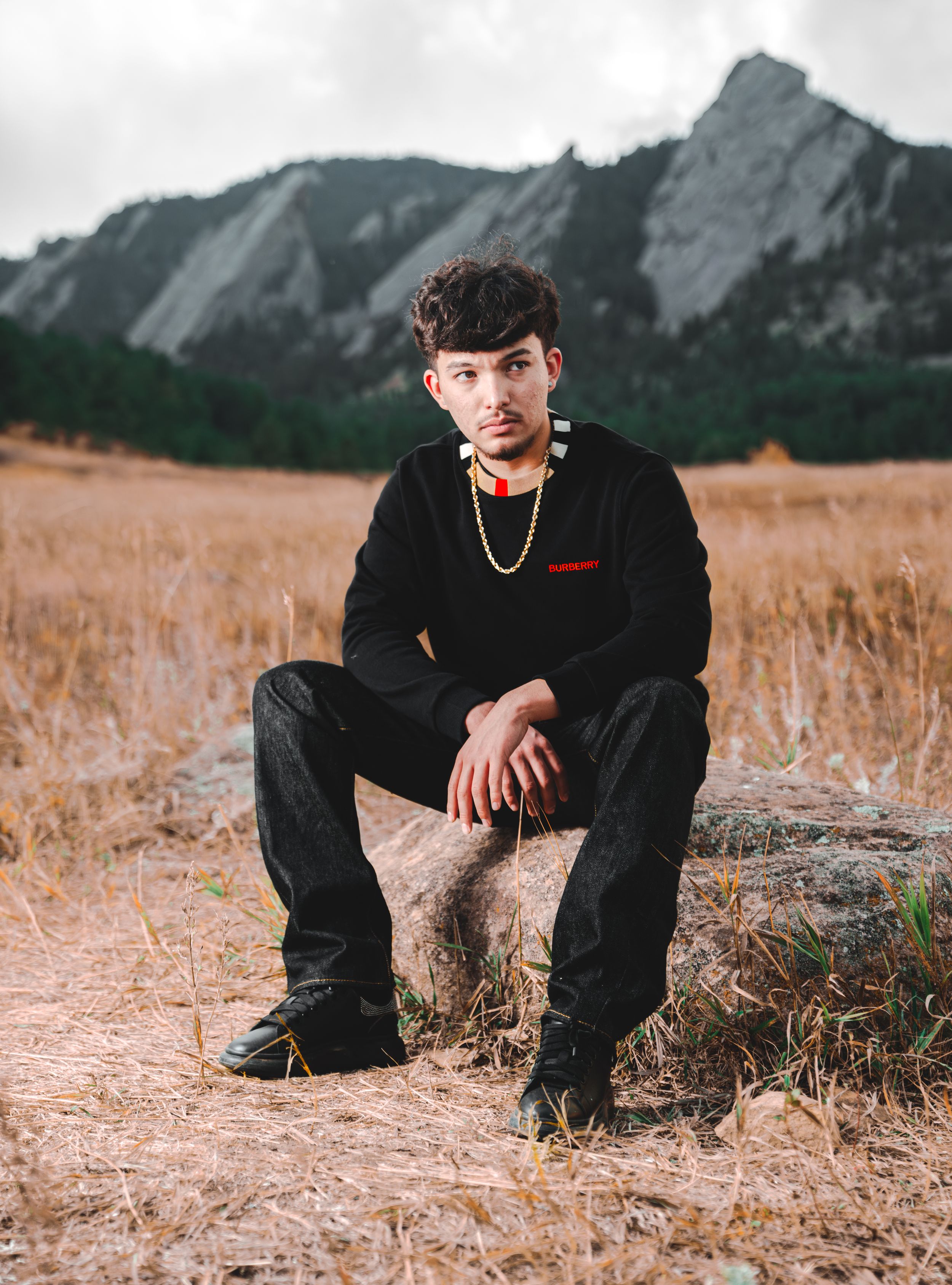 Person in dark clothing sits on ground against backdrop of dramatic mountain peaks in dry grass field at sunset.