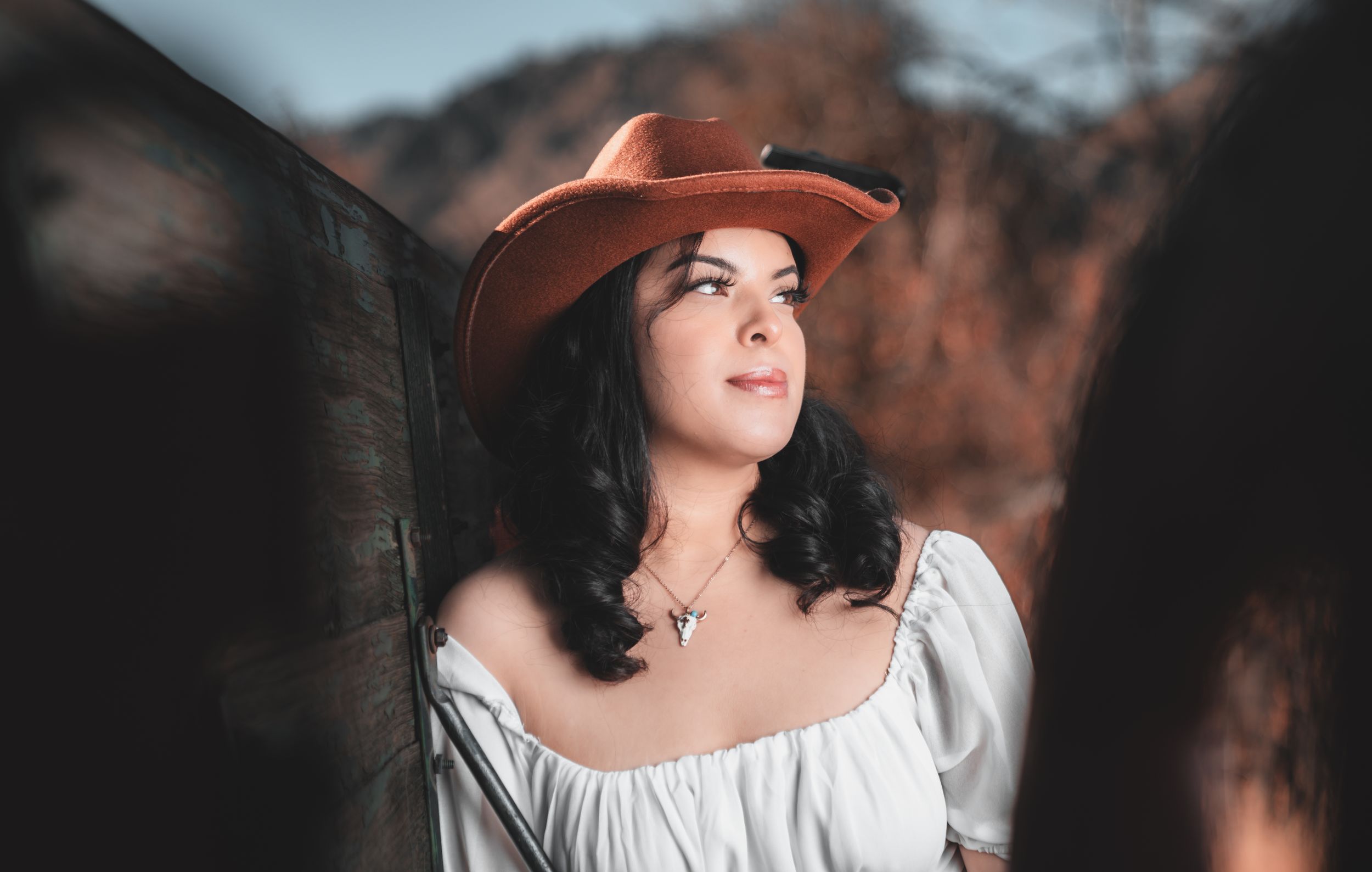 A person in a white peasant blouse and brown cowboy hat gazes upward against a dark rustic background.