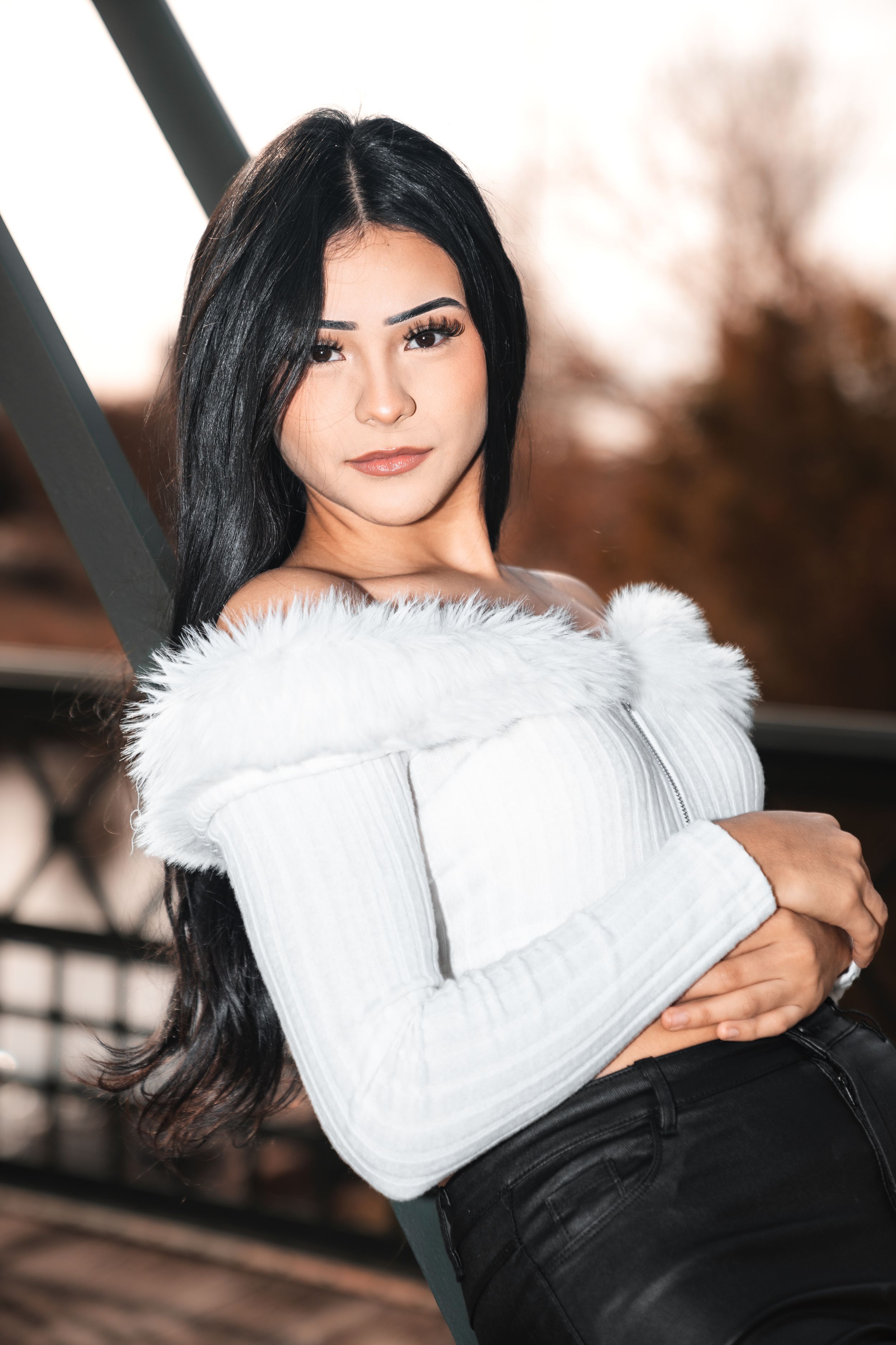 Portrait of a woman in a white fur-trimmed sweater and black pants posing outdoors against a blurred autumn background.