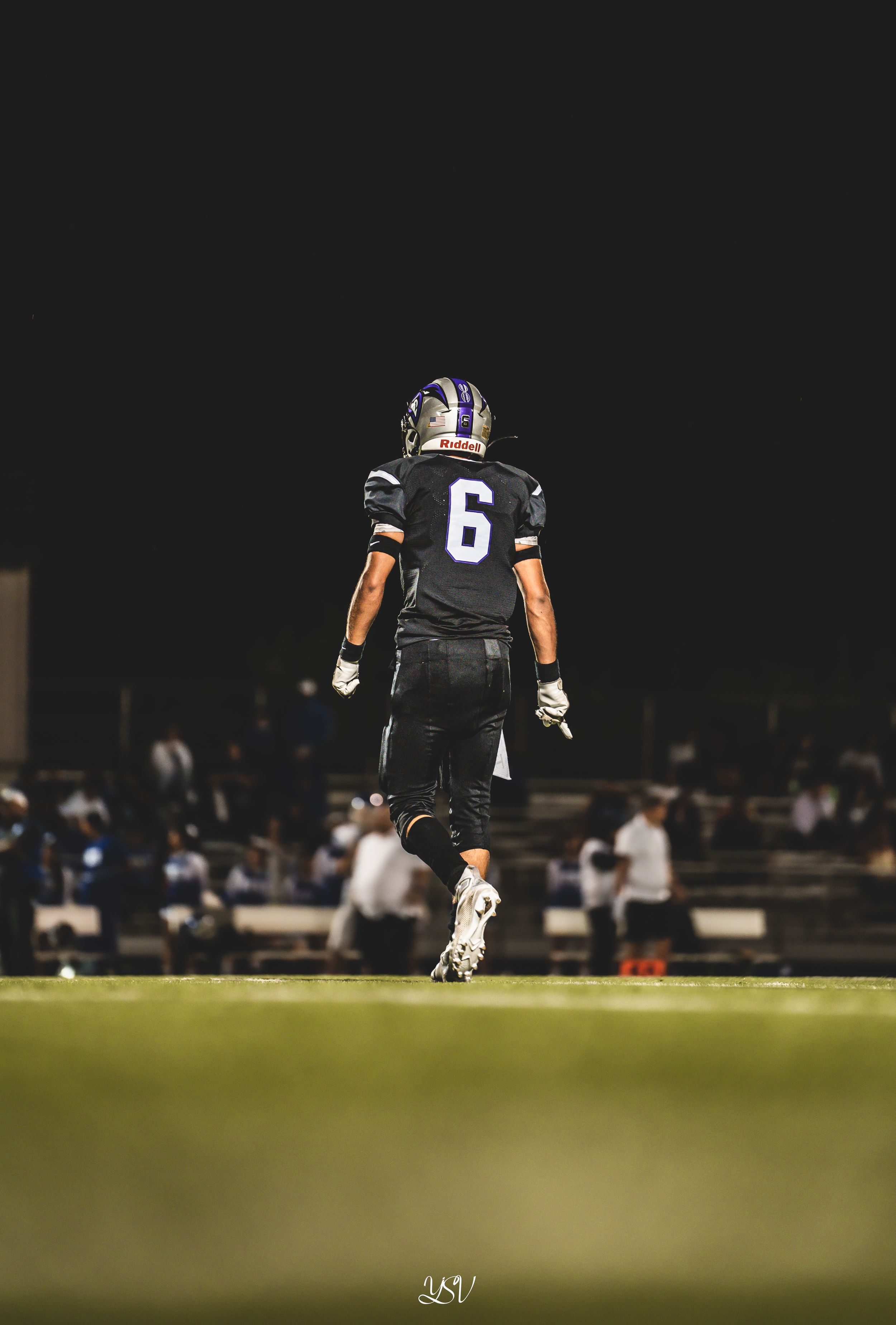 Football player wearing number 6 jersey stands on dark field during nighttime game under stadium lights.