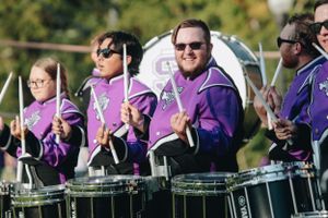 Marching band members play drums while wearing purple uniforms at an outdoor event.