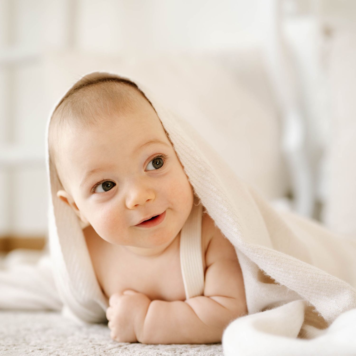 Sweet baby peeks out from under soft white blanket in bright nursery setting.