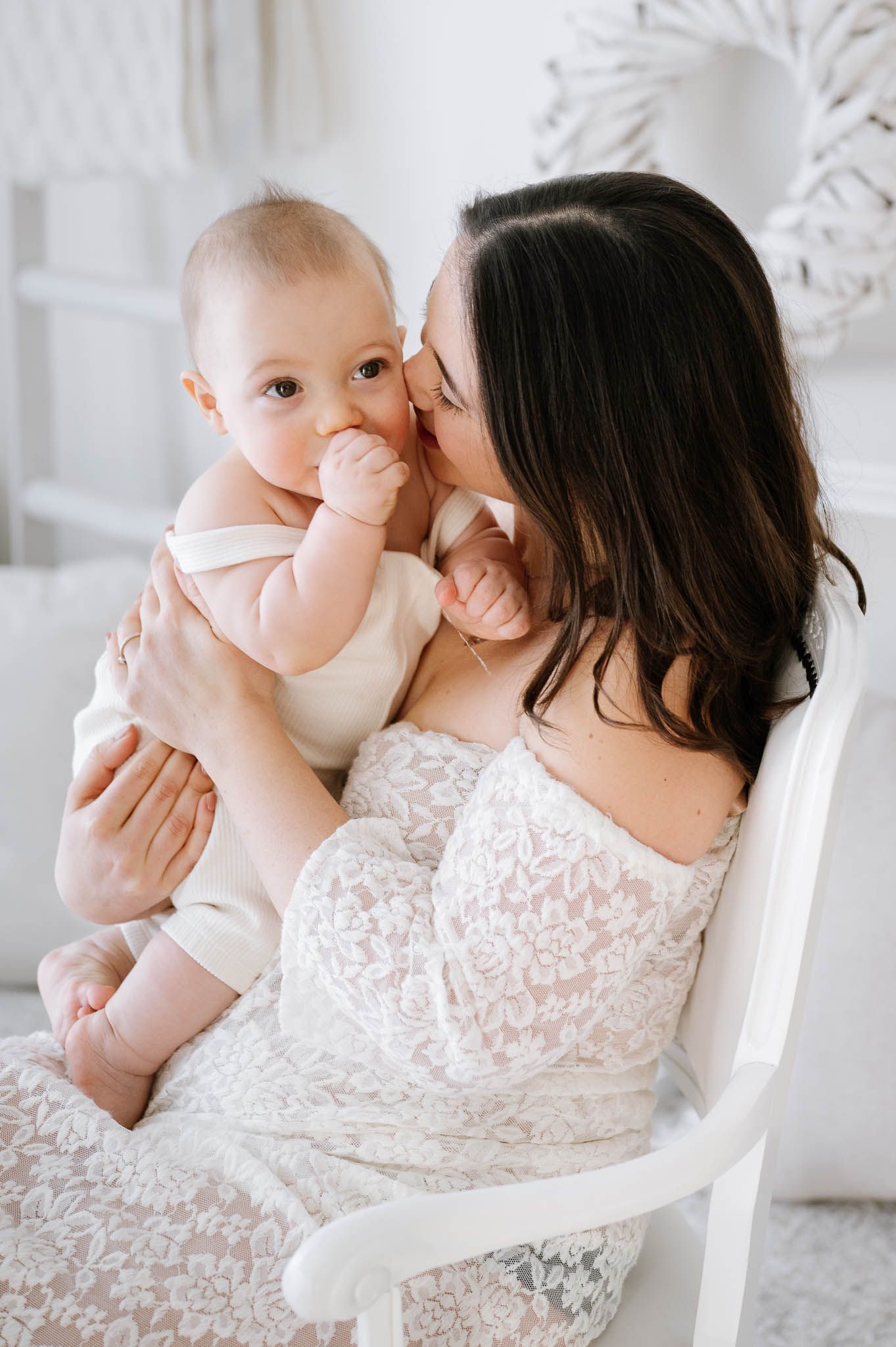 A woman in a white lace dress holds and kisses a baby in a bright, airy room with ornate white wall details.