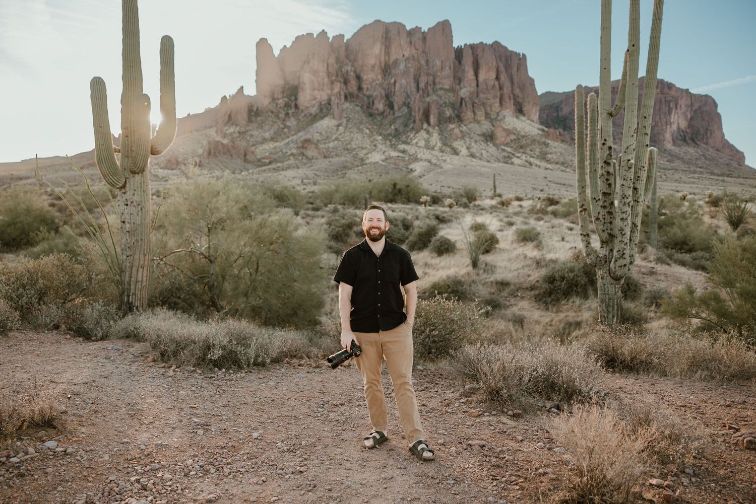 A photographer posed in front of the Superstition mountains nestled between two towering Saguaro cacti.