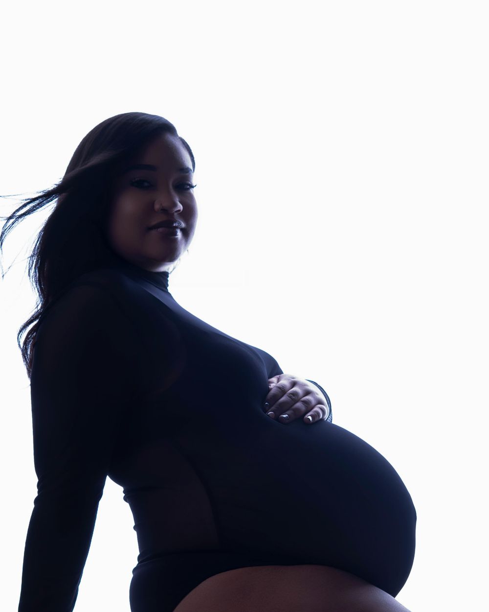 A silhouetted figure with flowing hair poses in a maternity portrait against a bright white backdrop.