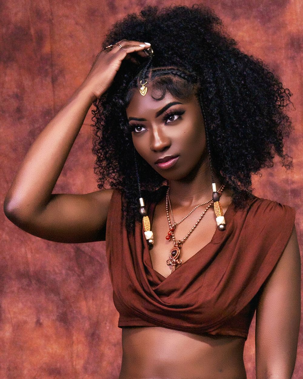 Beautiful portrait with curly natural hair wearing brown draped top and bohemian jewelry against textured rust background.