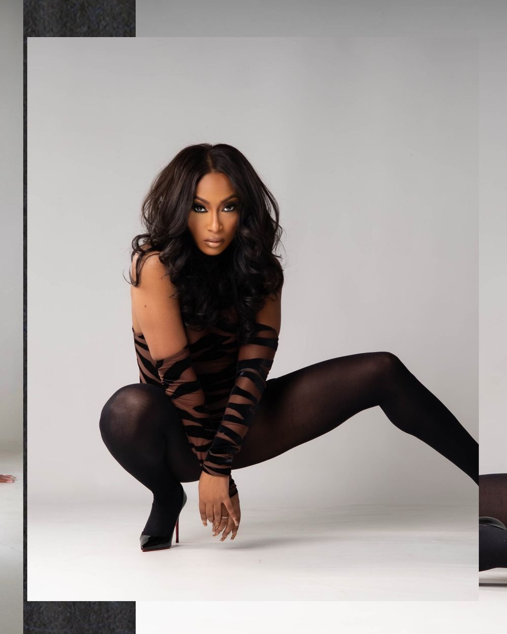Fashion model in black crouching pose against white studio background wearing artistic cut-out bodysuit and heels.