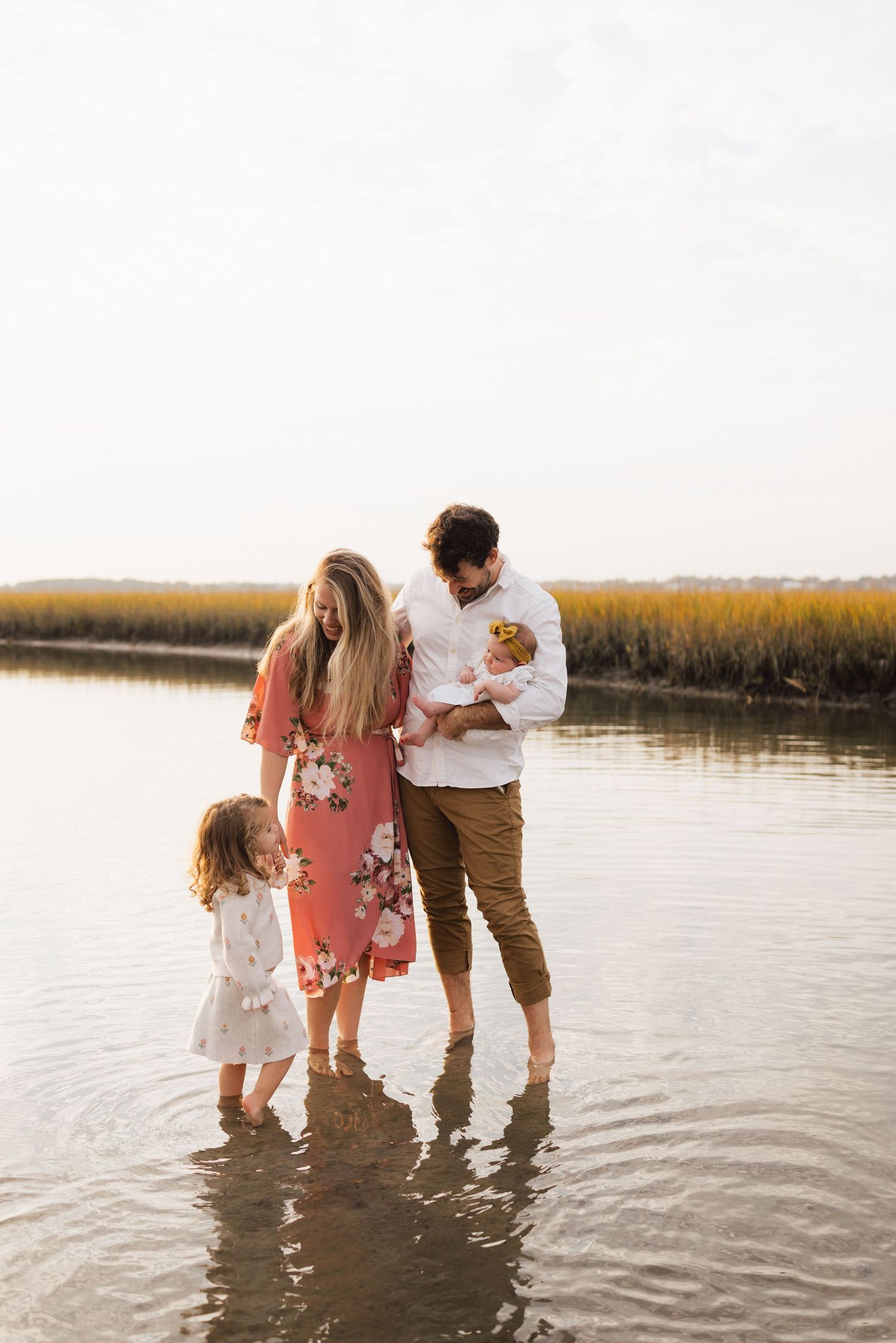 Family stands together in shallow coastal water at sunset with golden marsh grasses in background. Wilmington photographer.