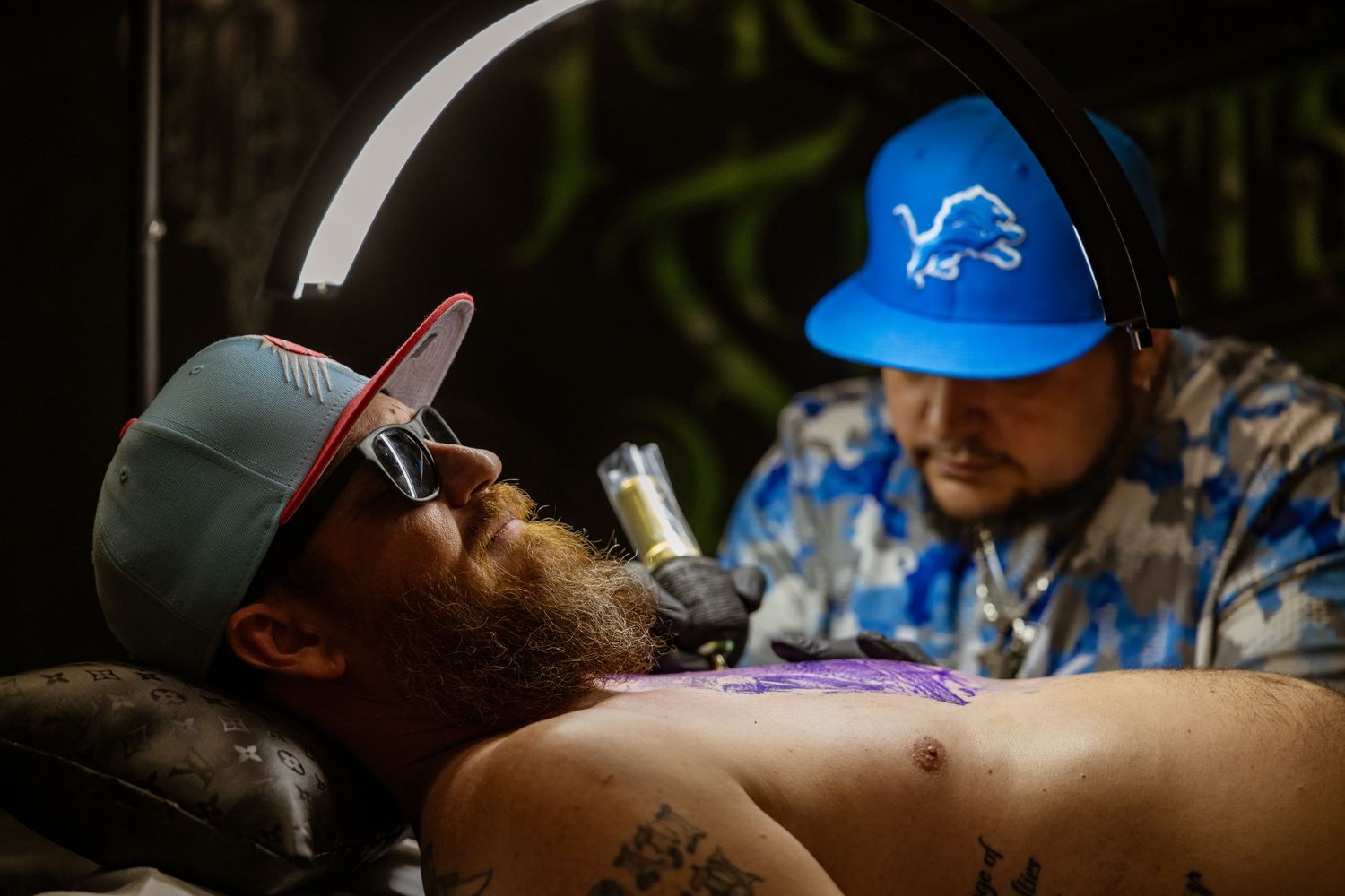 Artists at work in a tattoo studio with Detroit Lions caps and professional equipment.