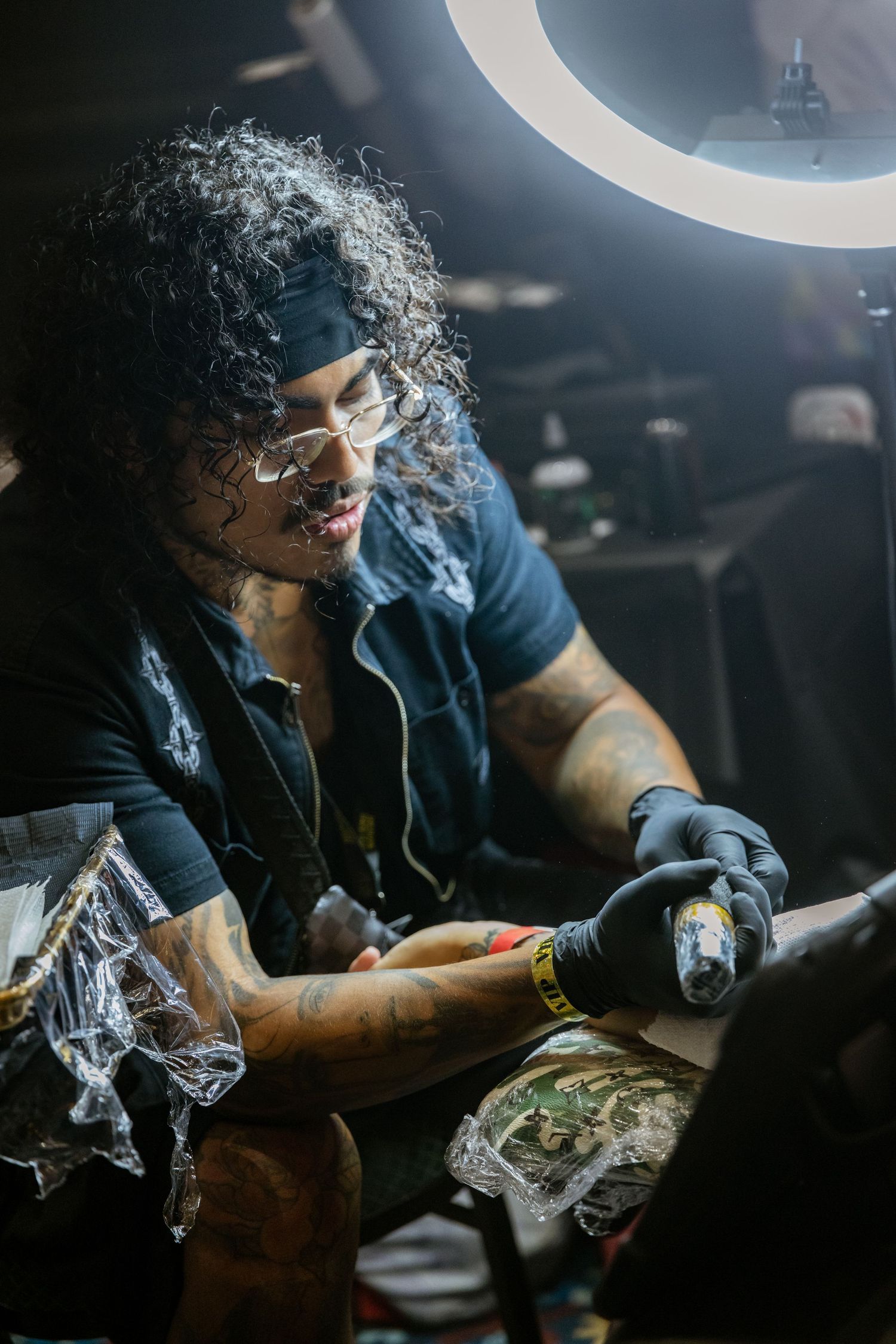 Tattoo artist working under bright ring light in a dark studio setting.