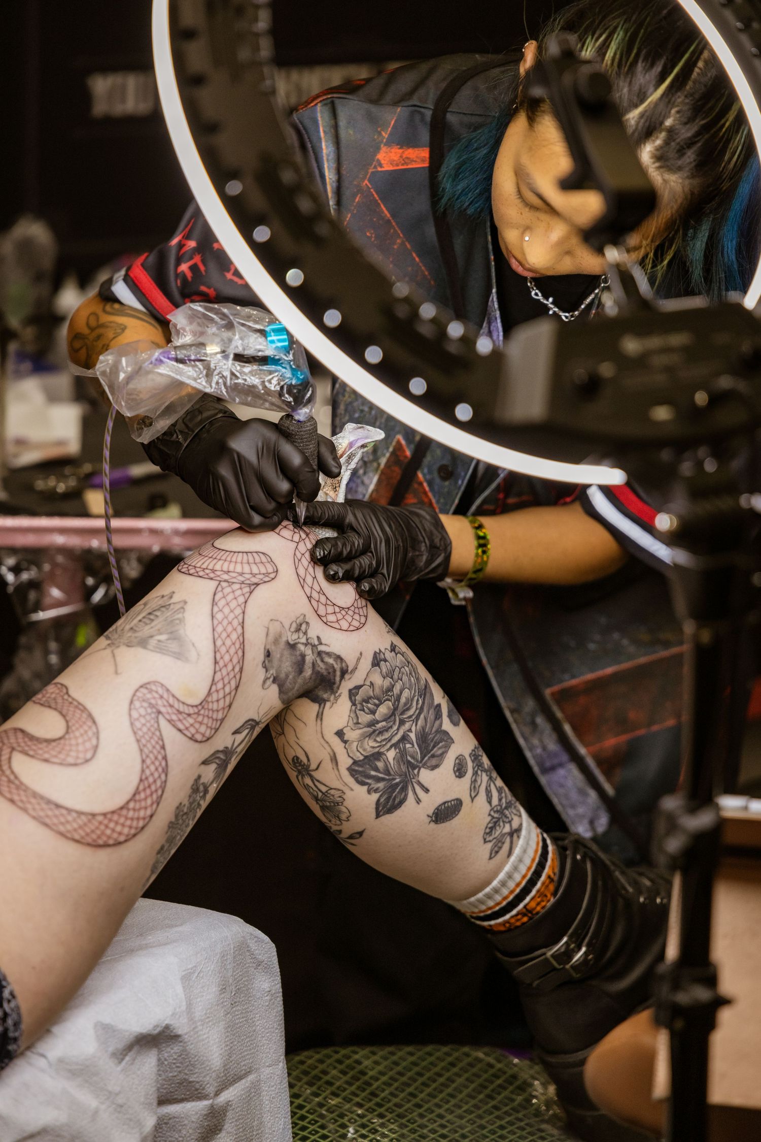 Artist wearing gloves working on leg tattoo under bright LED ring light.