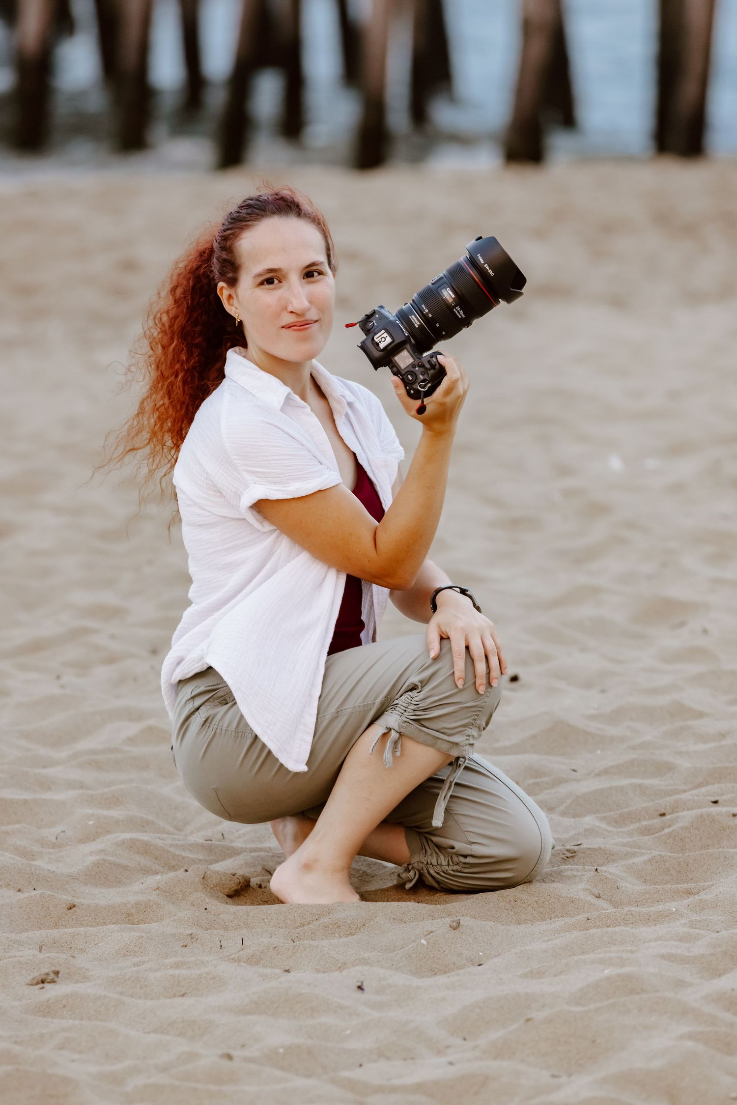 Female wedding photographer crouches on sandy beach holding professional camera near ocean pier. Virginia Beach.