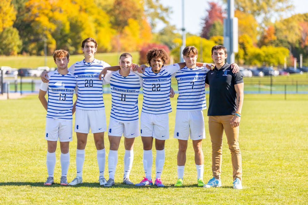 Soccer team poses in striped uniforms on sunny autumn field with fall foliage in background.
