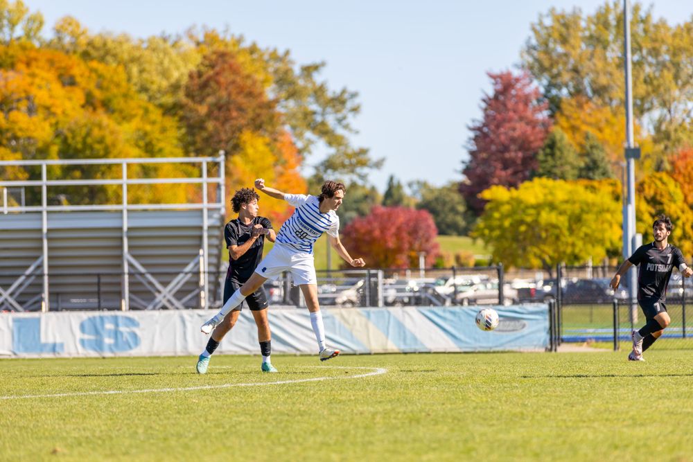Soccer match in progress on grass field with autumn trees and bleachers in background.