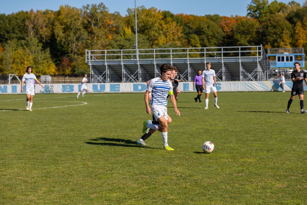 Soccer players compete on grass field with fall foliage and sports facilities visible.