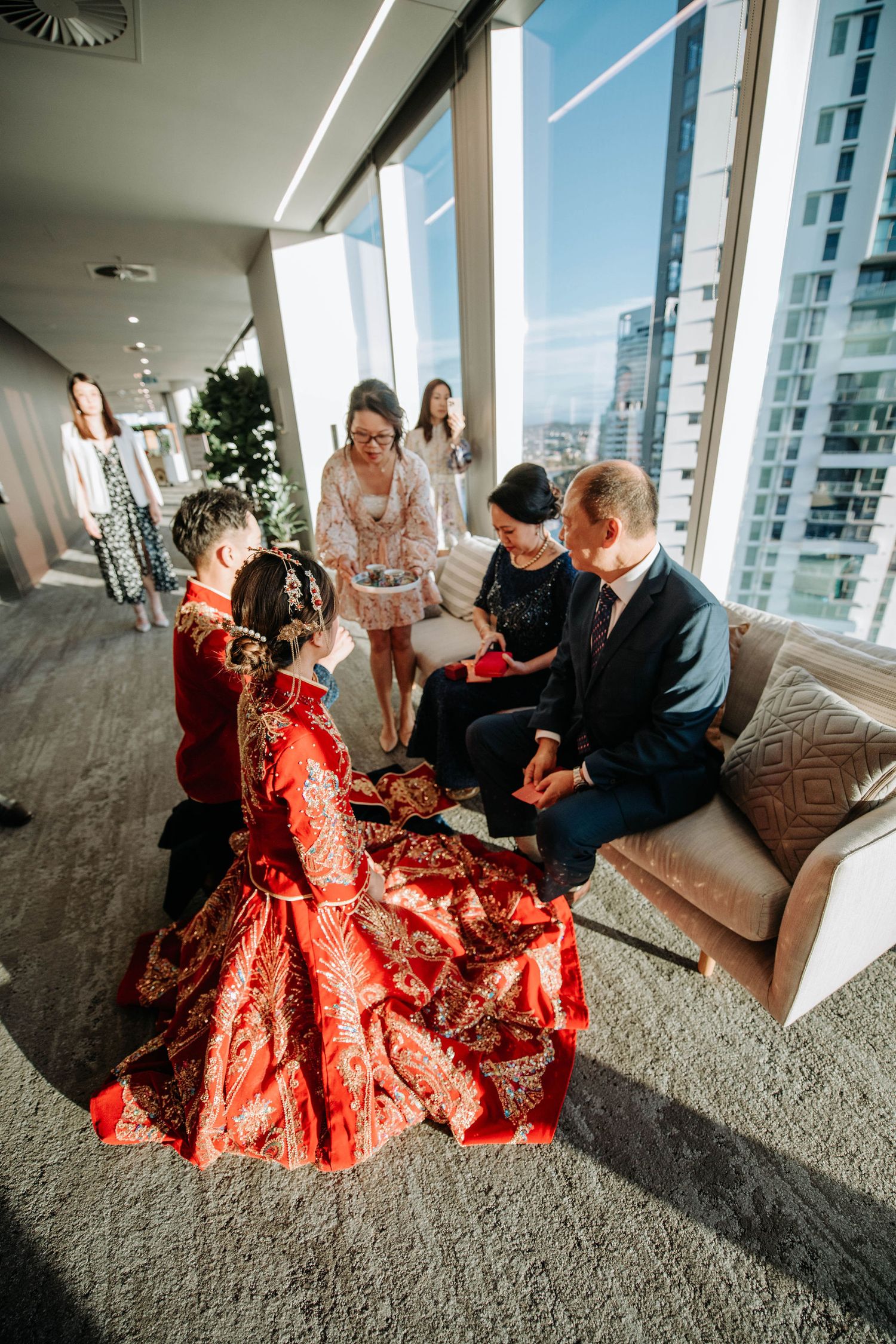A wedding tea ceremony with the couple in traditional red Chinese attire in a modern high-rise setting.