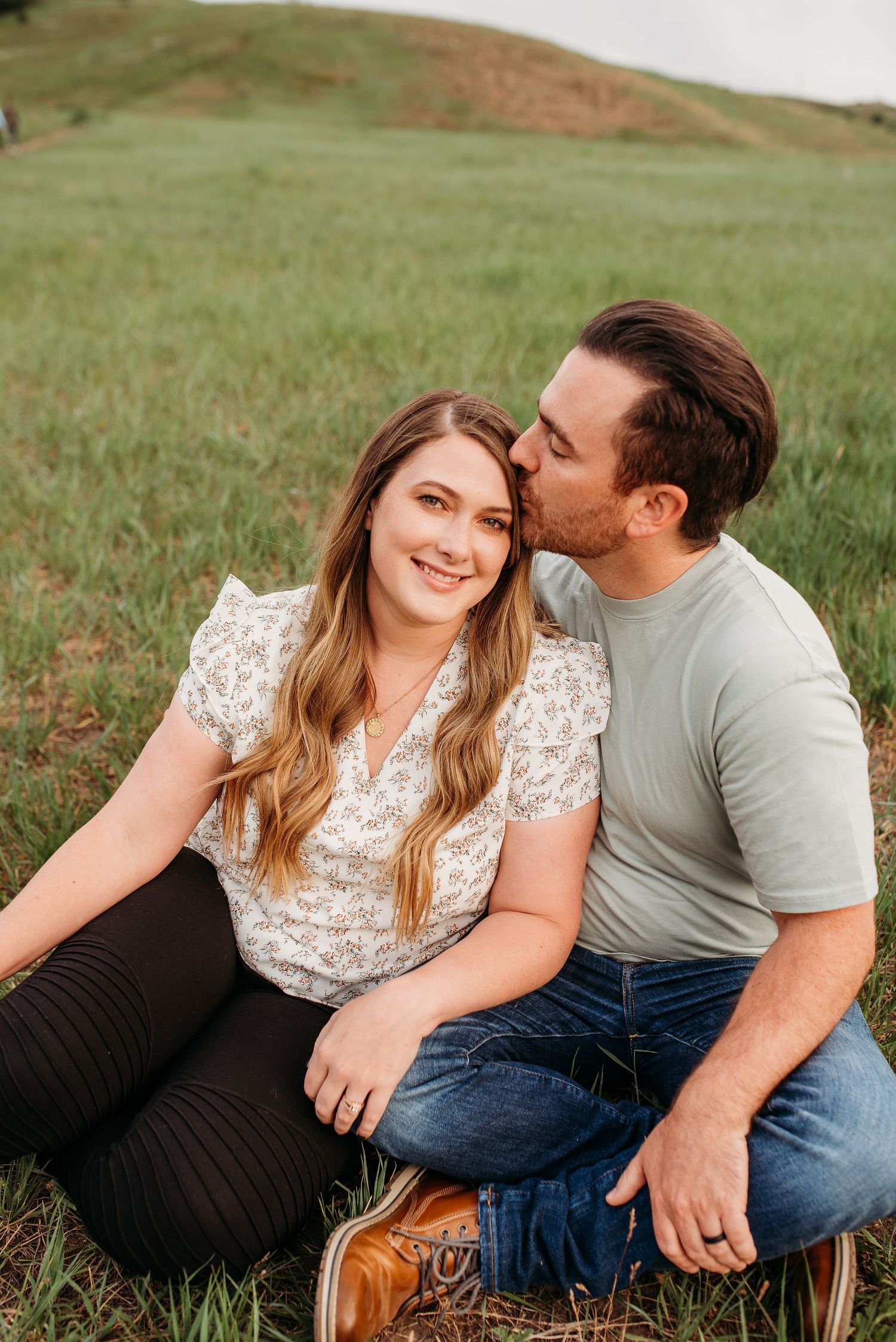 A loving couple sits together on grassy hillside during golden hour.