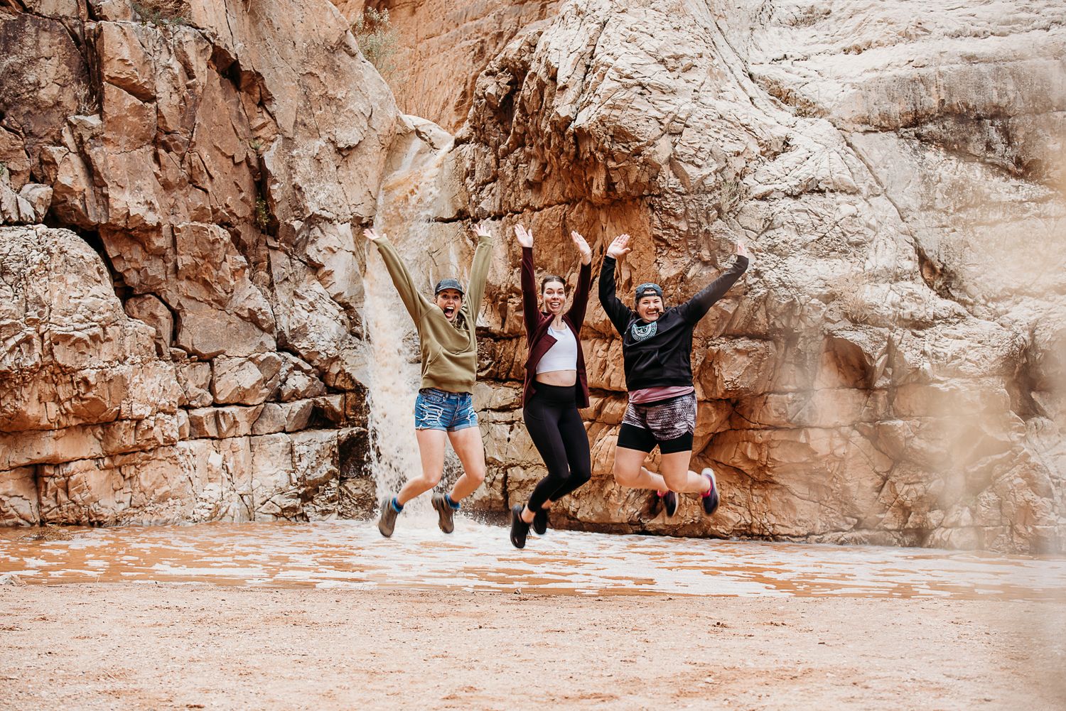A group of friends jumping joyfully in front of red rock formations in an outdoor canyon setting.