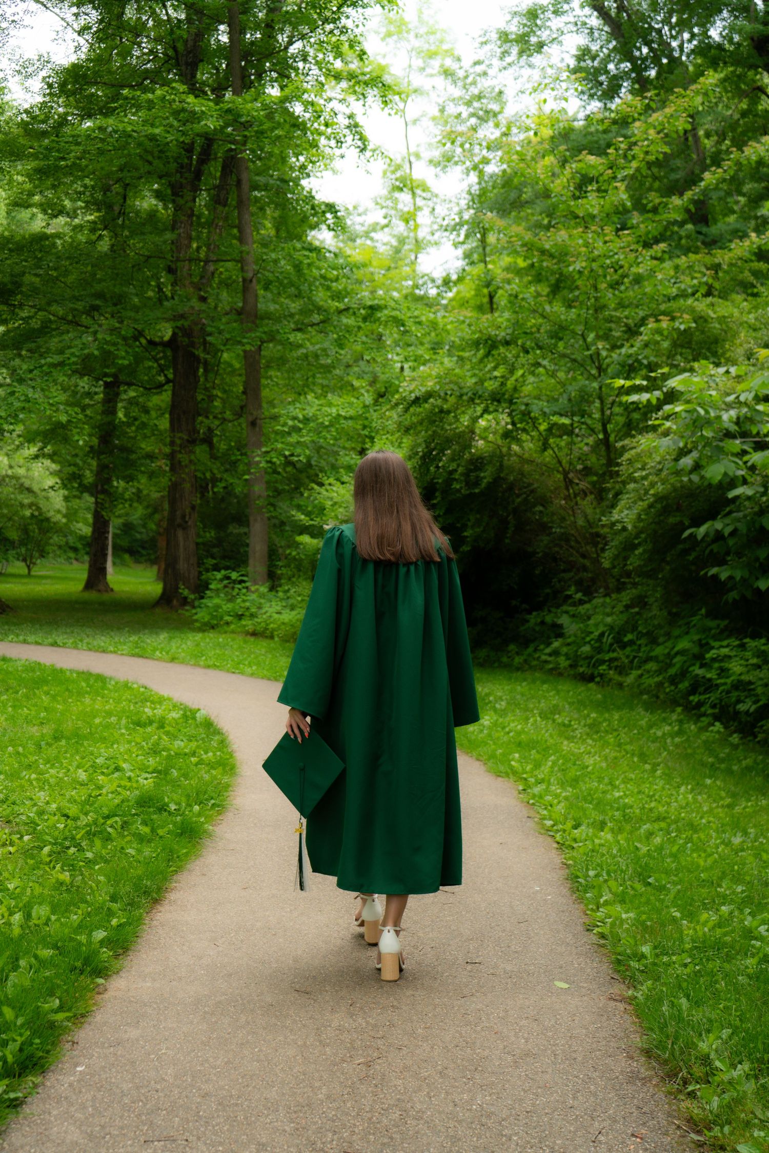 Someone in emerald green graduation gown walks alone along curved path through lush summer park.