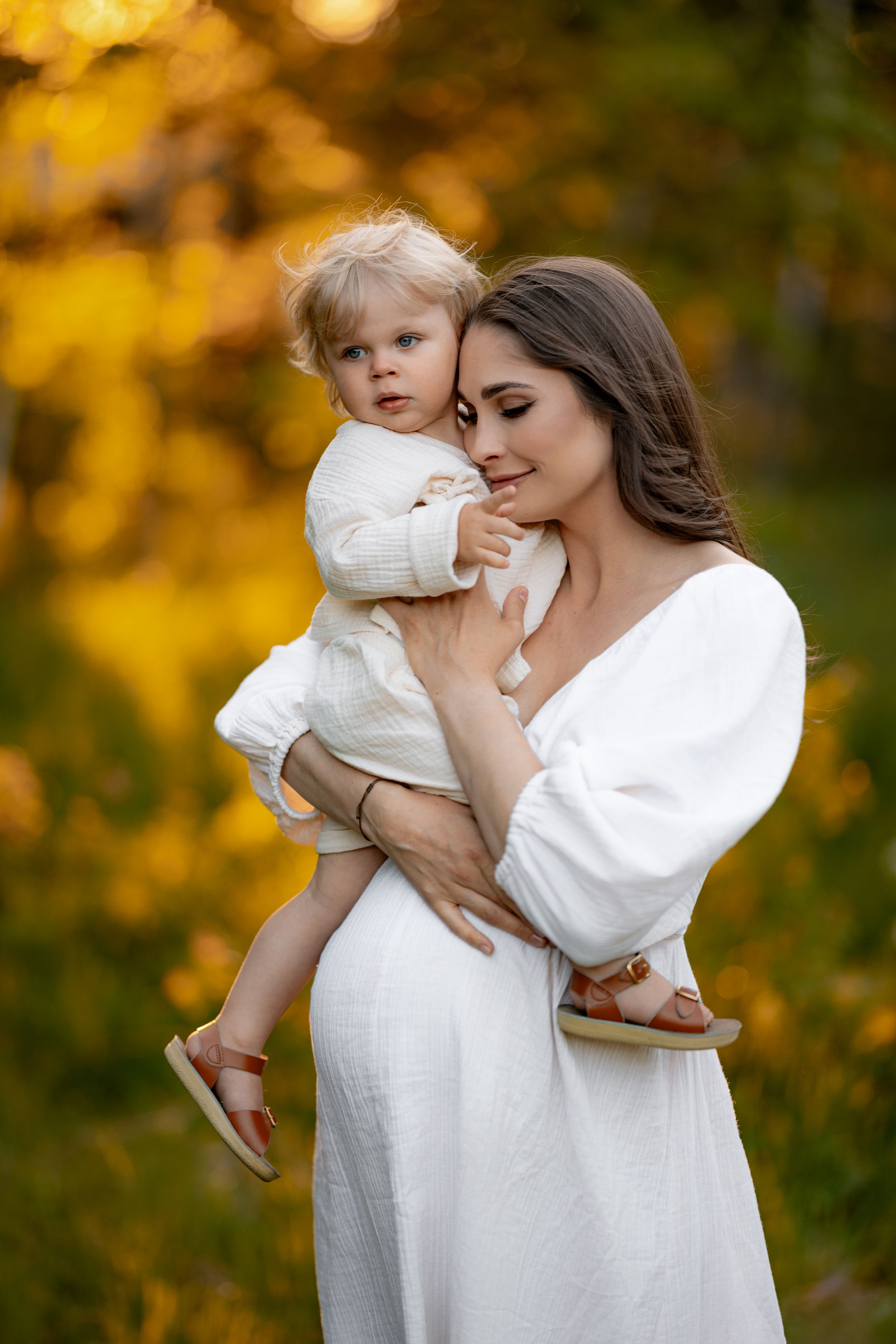 A woman in a white dress holds a blonde toddler against autumn trees.