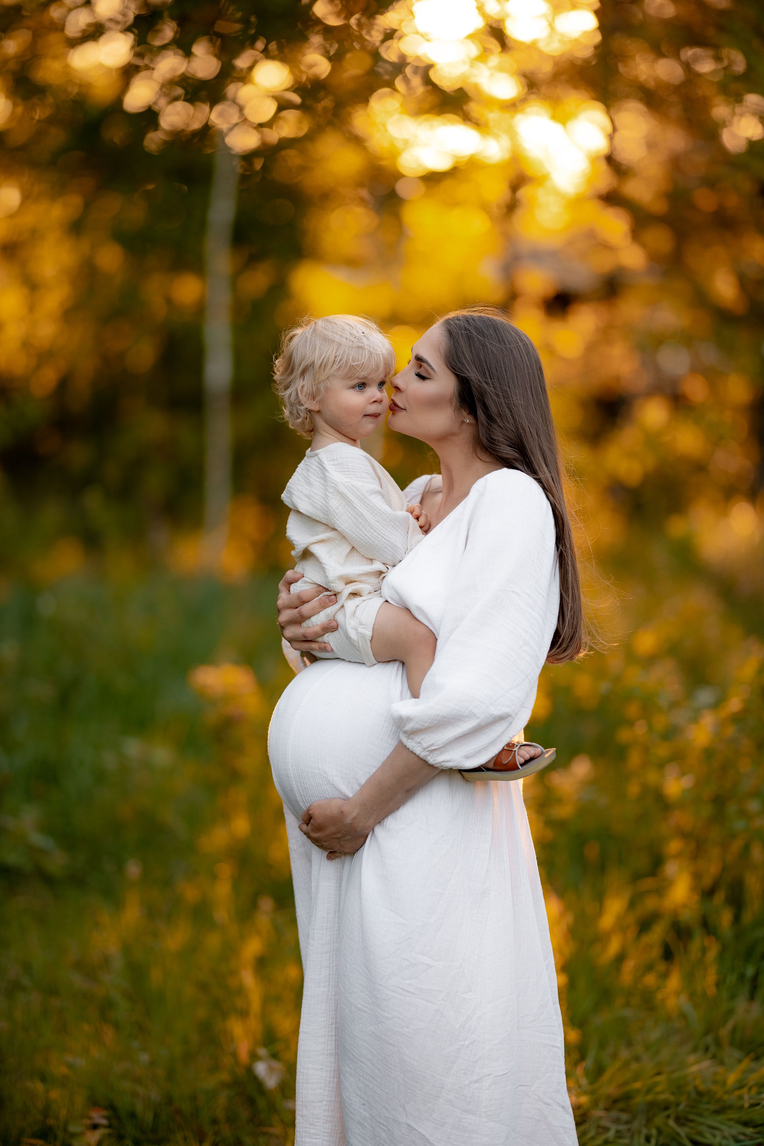 A woman in a flowing white dress holds a small child amid autumn foliage.