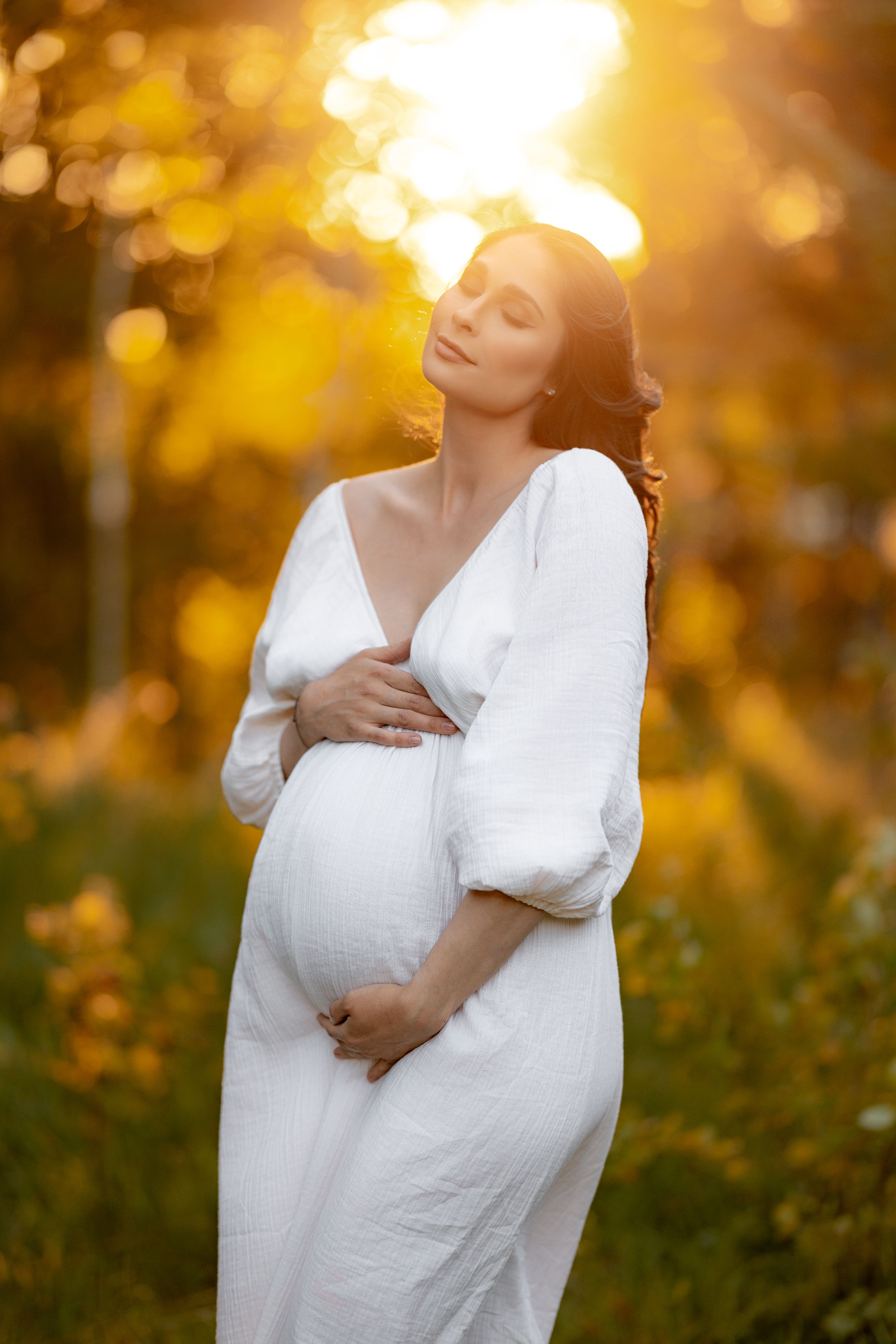 A woman in a white dress cradles her pregnant belly in golden sunlight.