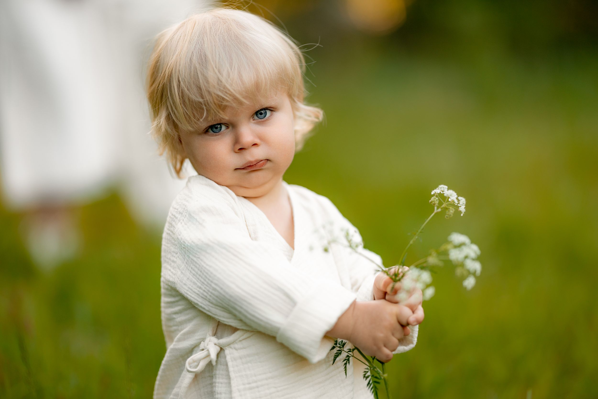 Young child in white holding dandelions in a lush green setting.