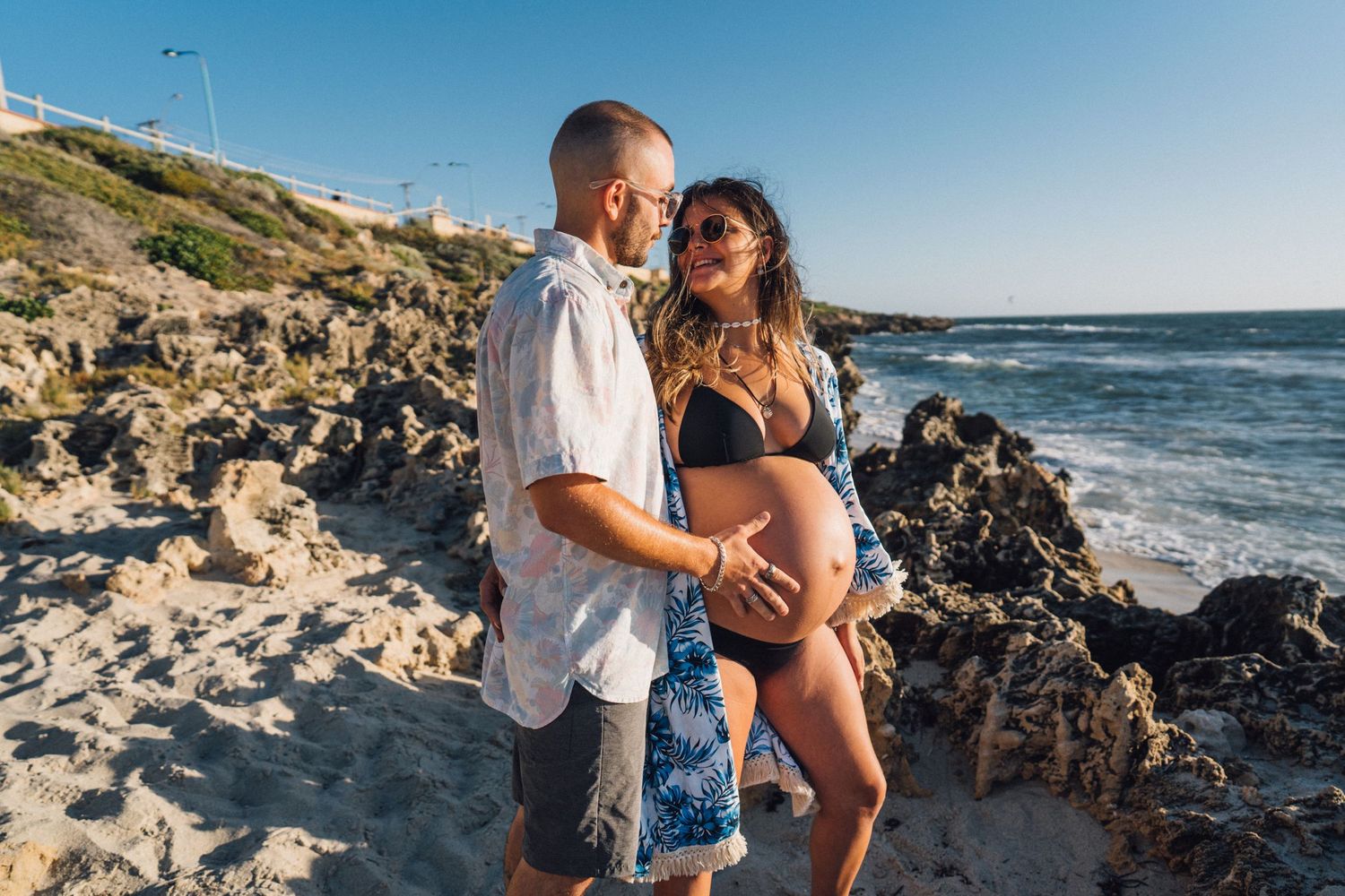 A couple shares an intimate moment on a rocky beach during sunset against a dramatic ocean backdrop.