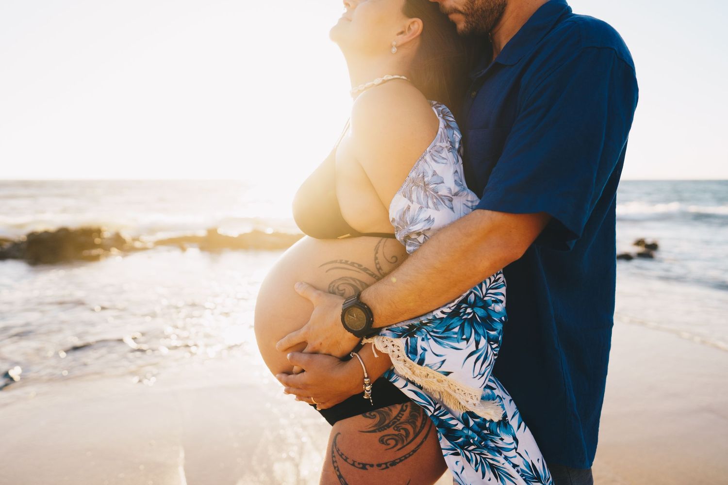 A tender maternity moment captured on the beach during golden hour with ocean waves behind.