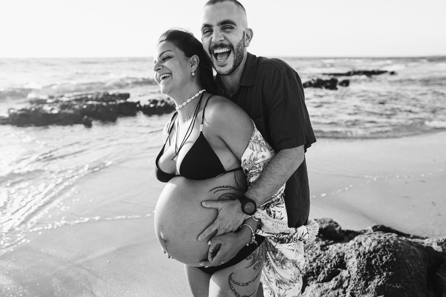 A joyful maternity photo session on a beach with rocky coastline and ocean waves.