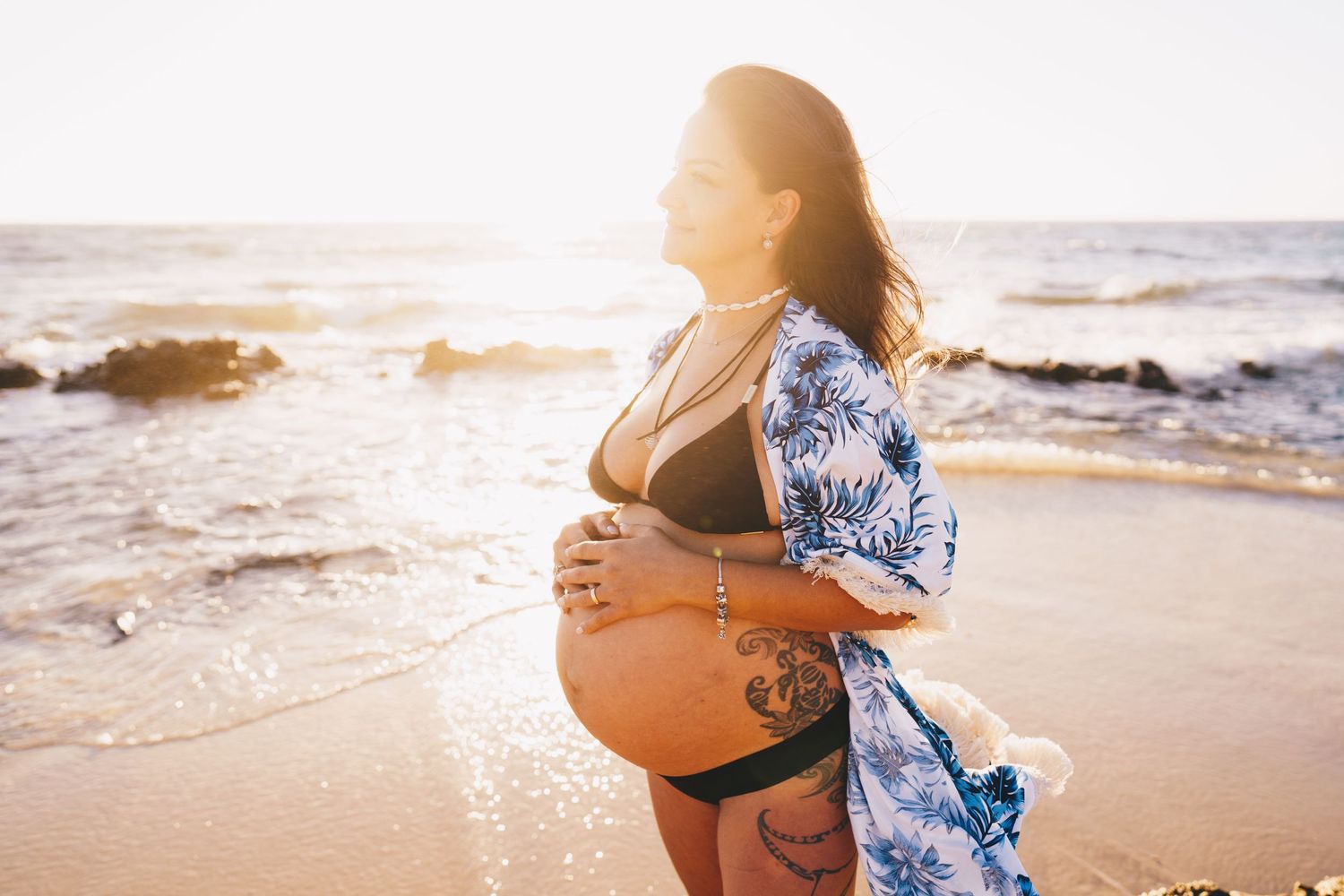 A maternity photoshoot at sunset on a beach with blue floral kimono and black bikini.