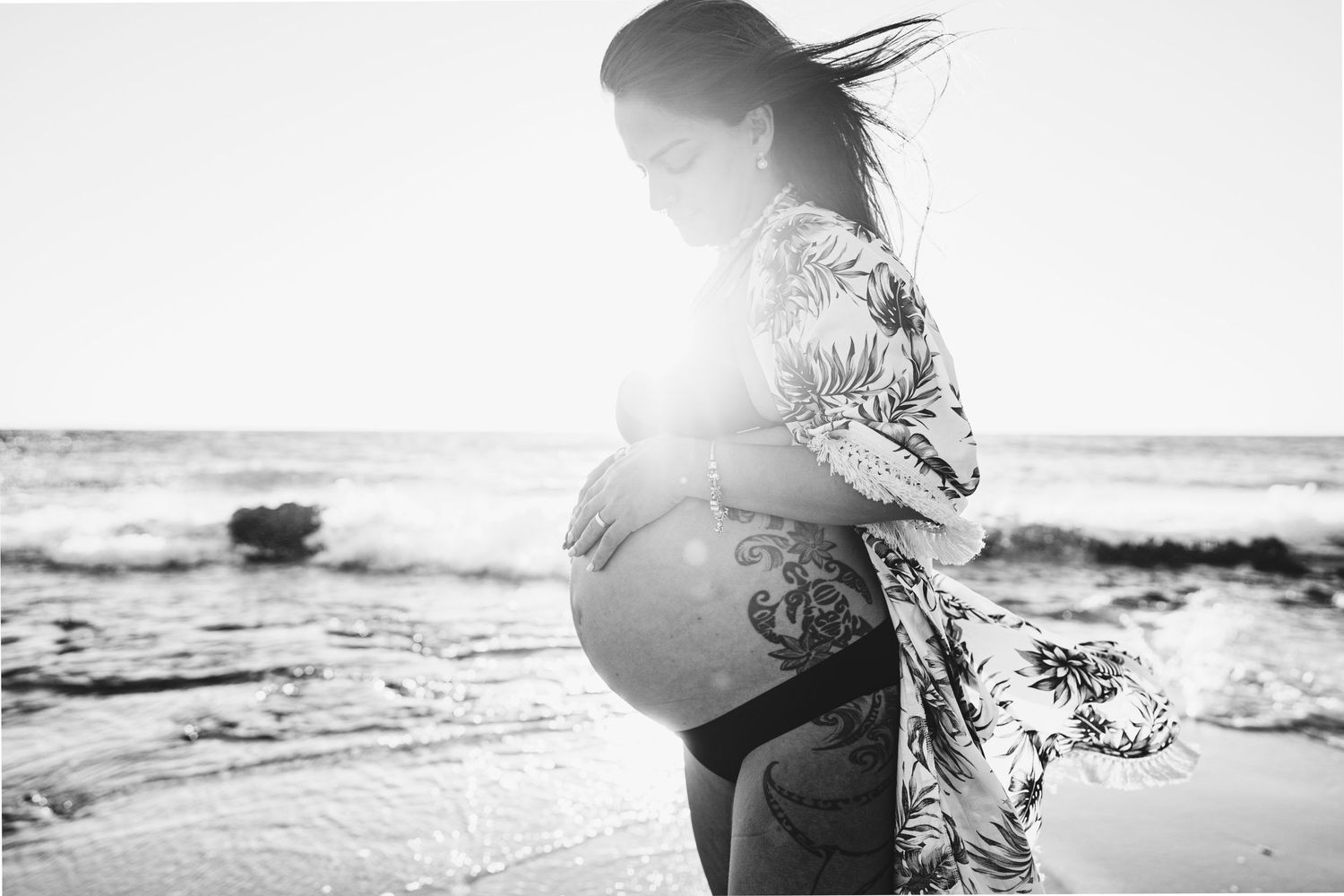 A black and white maternity portrait with sunlight flare on a rocky beach.