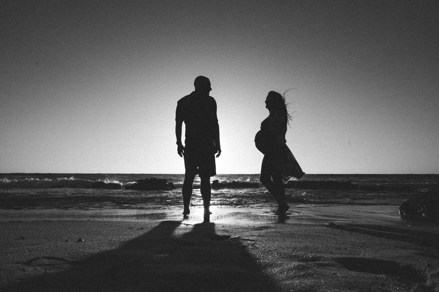 Black and white beach photo of two silhouettes against setting sun.