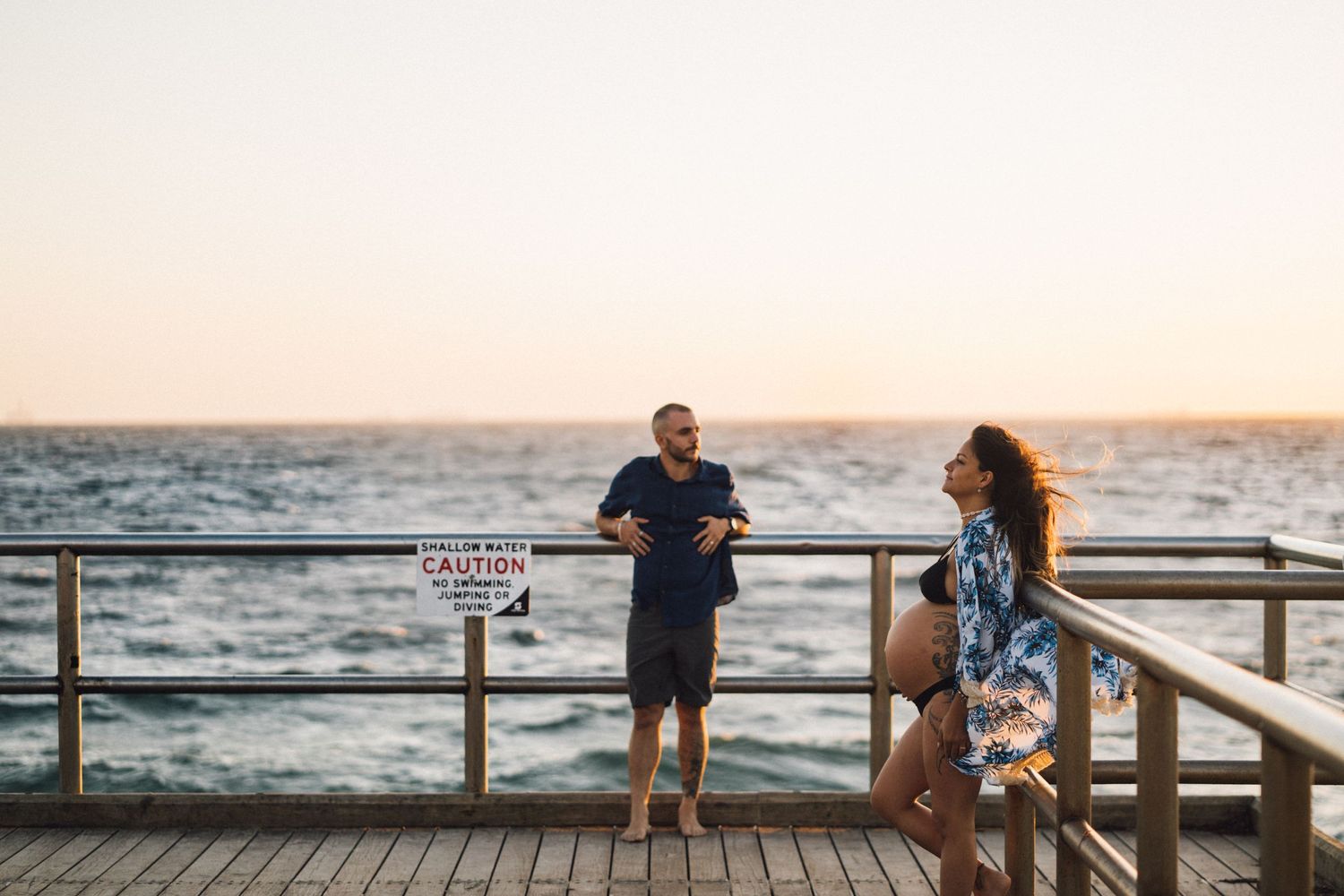 Couple standing on oceanfront boardwalk during sunset with warning sign visible.