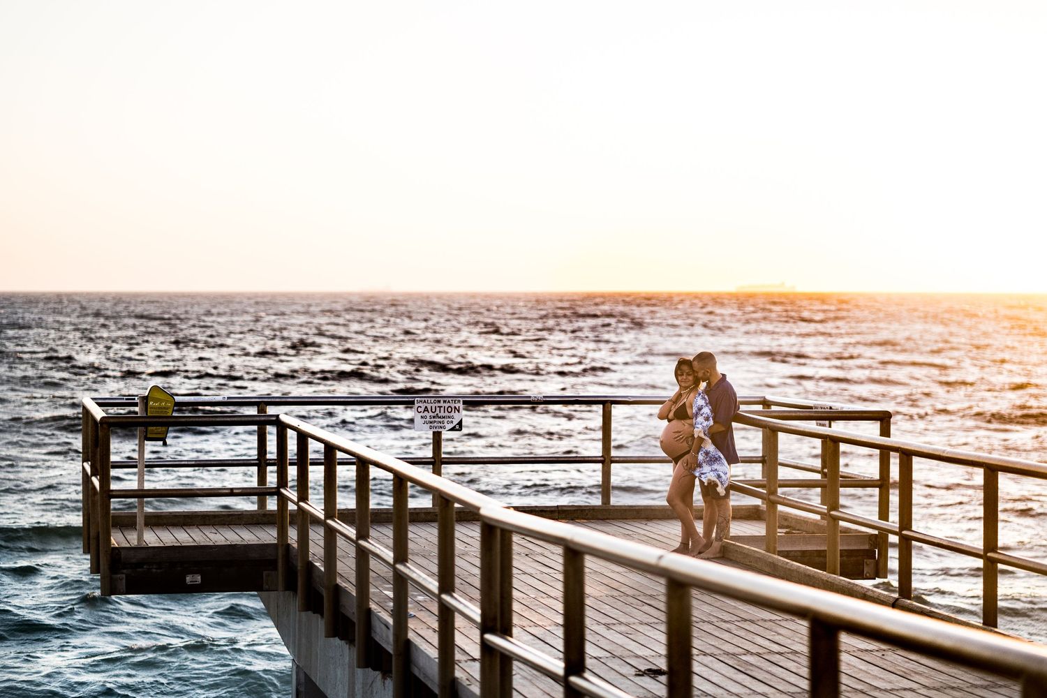 A couple shares an intimate moment at sunset on a wooden pier overlooking the ocean.