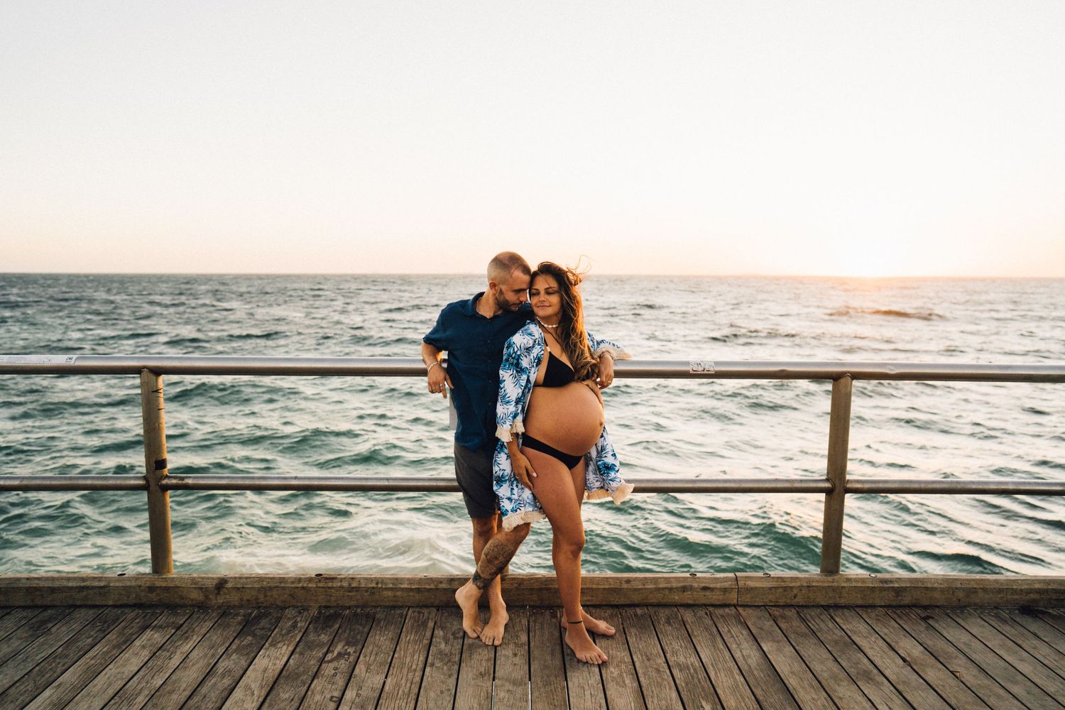 Expecting parents embrace on a wooden pier during golden hour with waves crashing behind them.