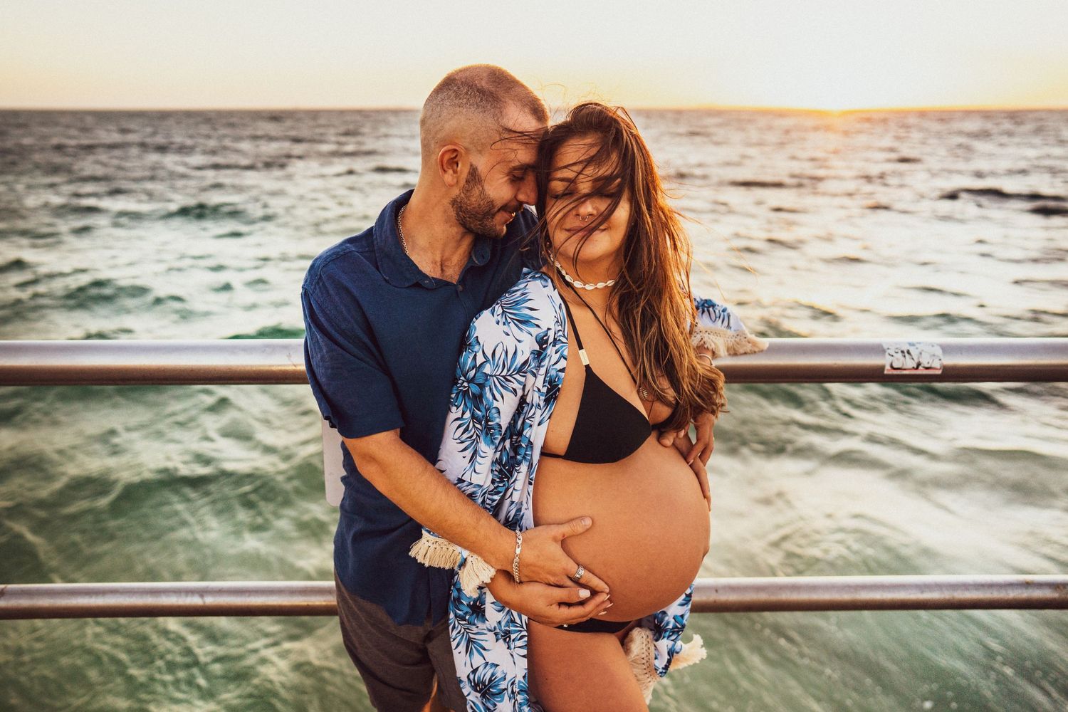 Intimate maternity photo of couple on pier during sunset with ocean waves in background.