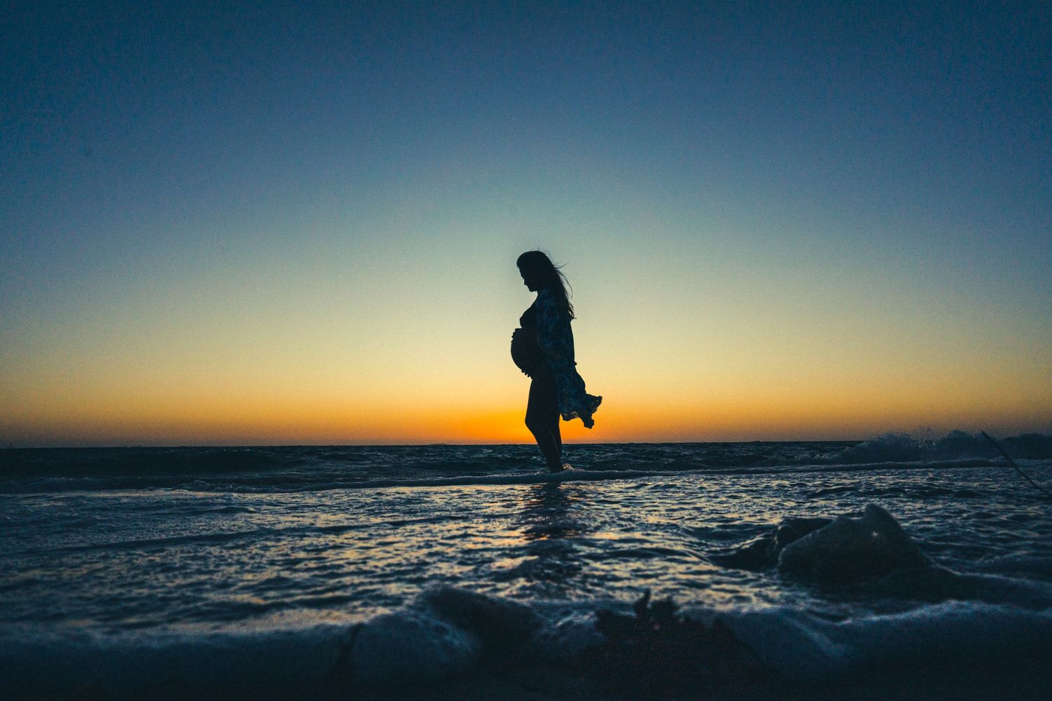 Silhouette of pregnant woman standing in ocean water during dramatic sunset.