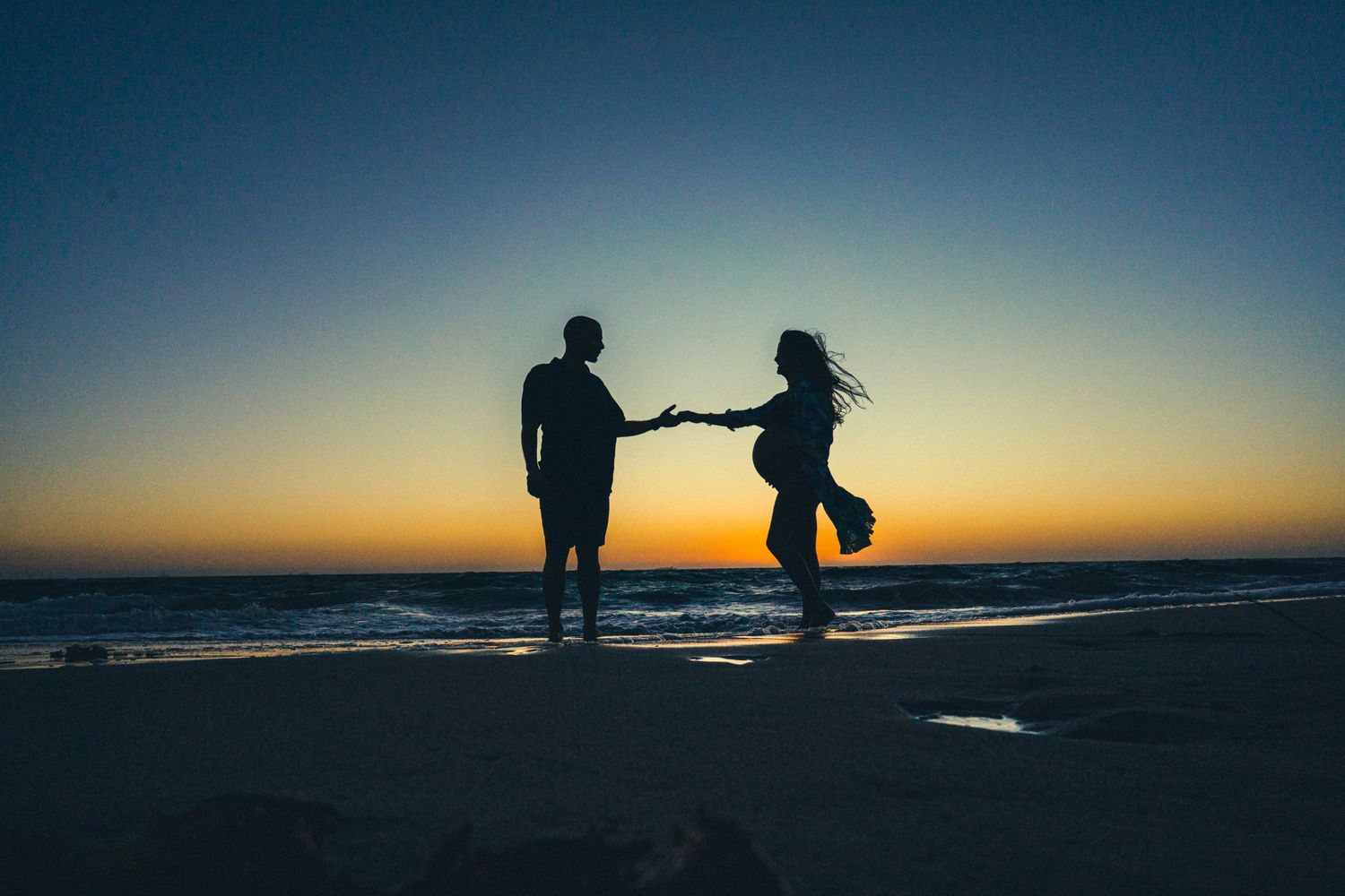 Silhouetted couple holds hands at beach during sunset with ocean waves in background.