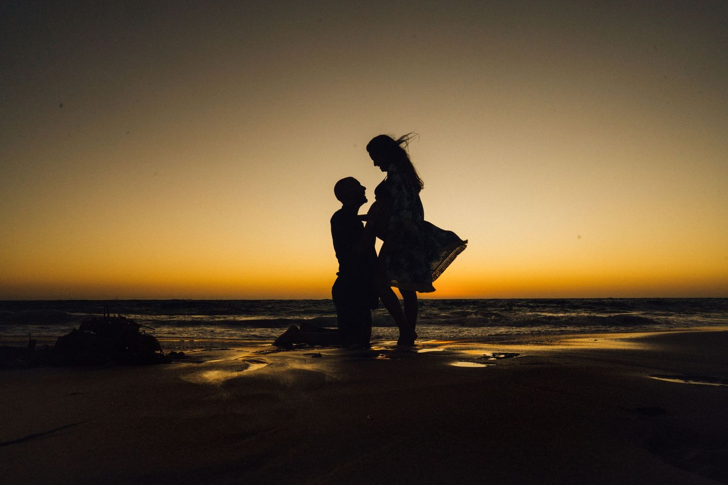 Silhouette of couple in romantic pose against golden sunset over beach waters.
