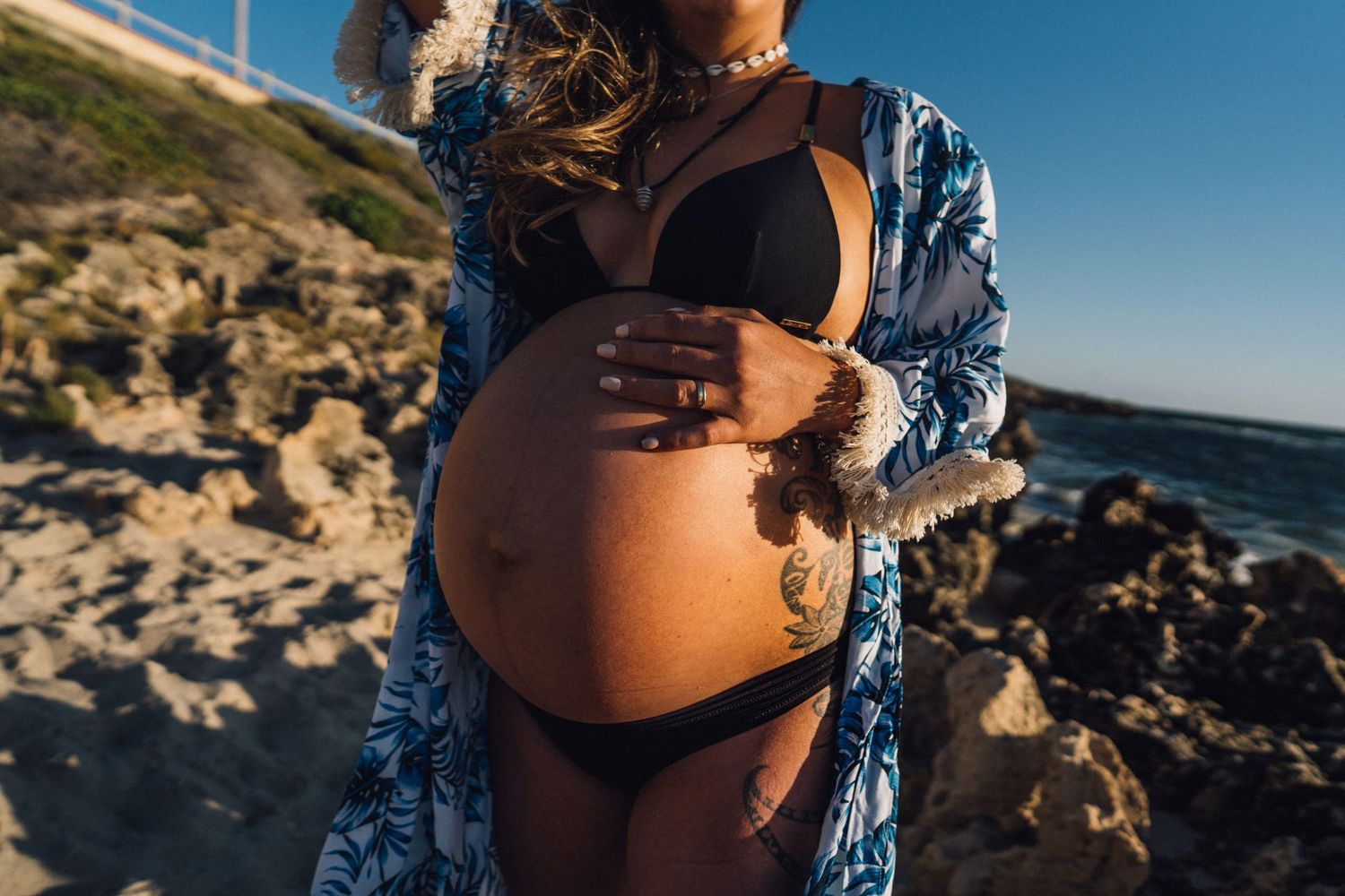 An expectant mother in a blue robe poses on rocky coastline with waves crashing in background.