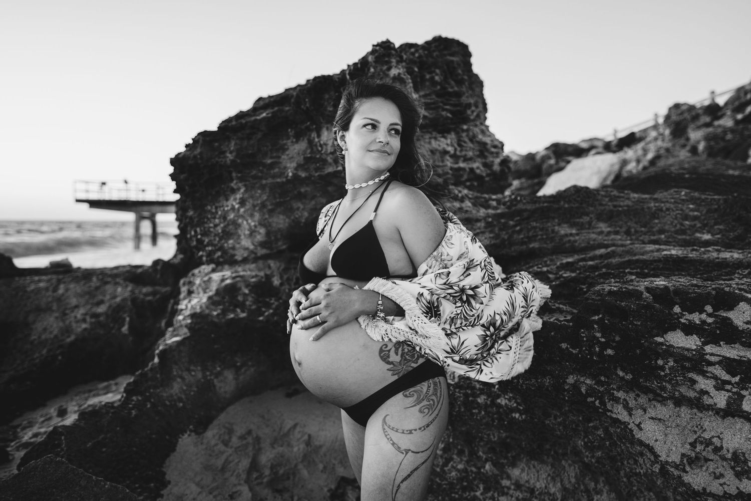 Black and white maternity portrait of woman in swimwear posed against coastal rocks.