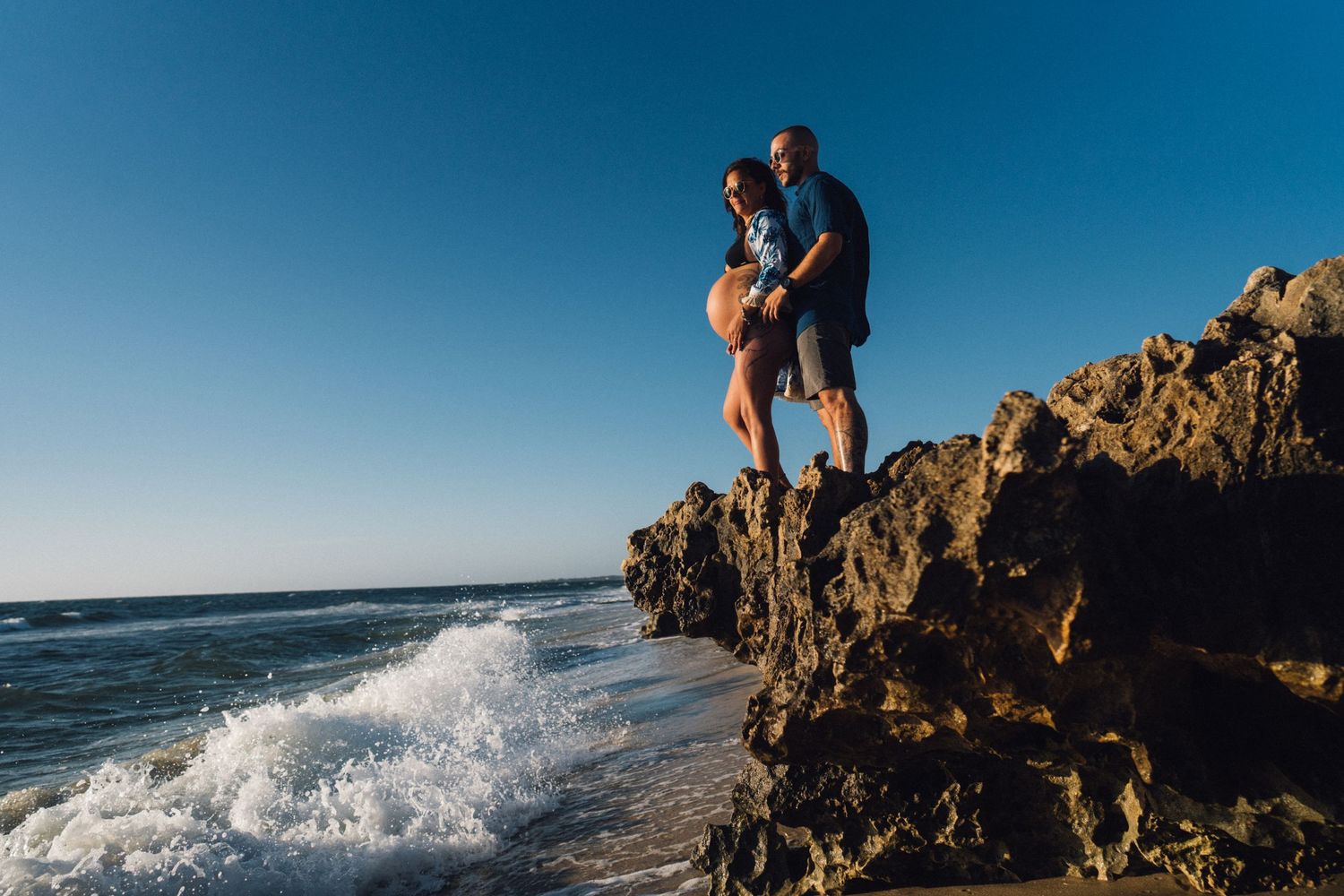 A couple stands atop coastal rocks while waves crash dramatically below them at the beach.