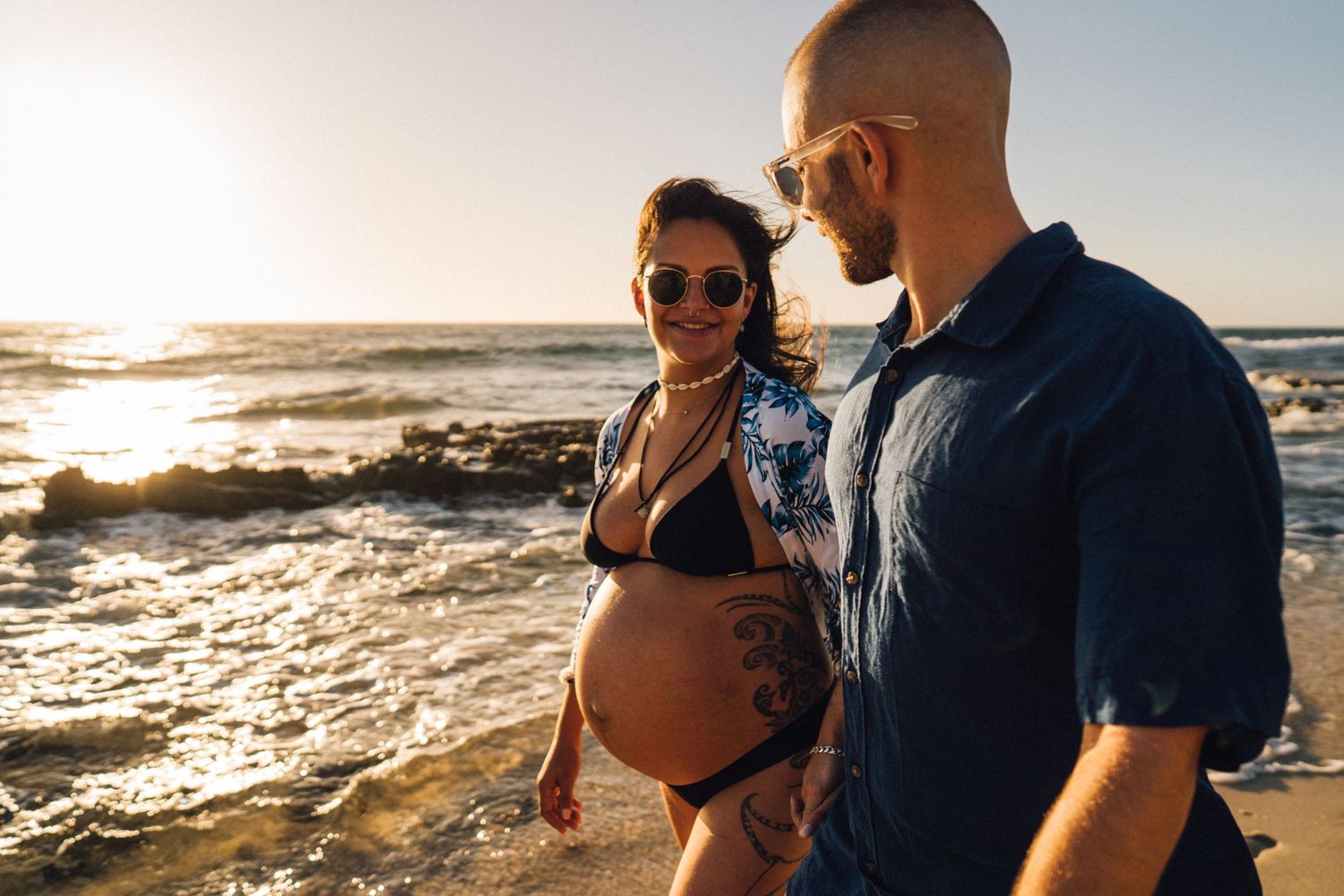 A romantic beachside maternity photo during golden hour with crashing waves and rocky coastline.