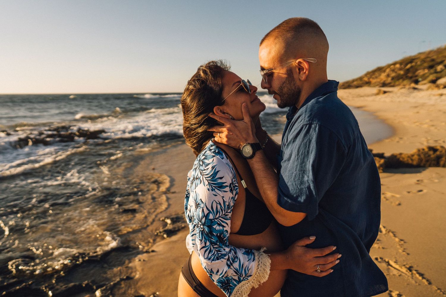 A tender moment between a couple on a sandy beach at sunset with ocean waves in background.
