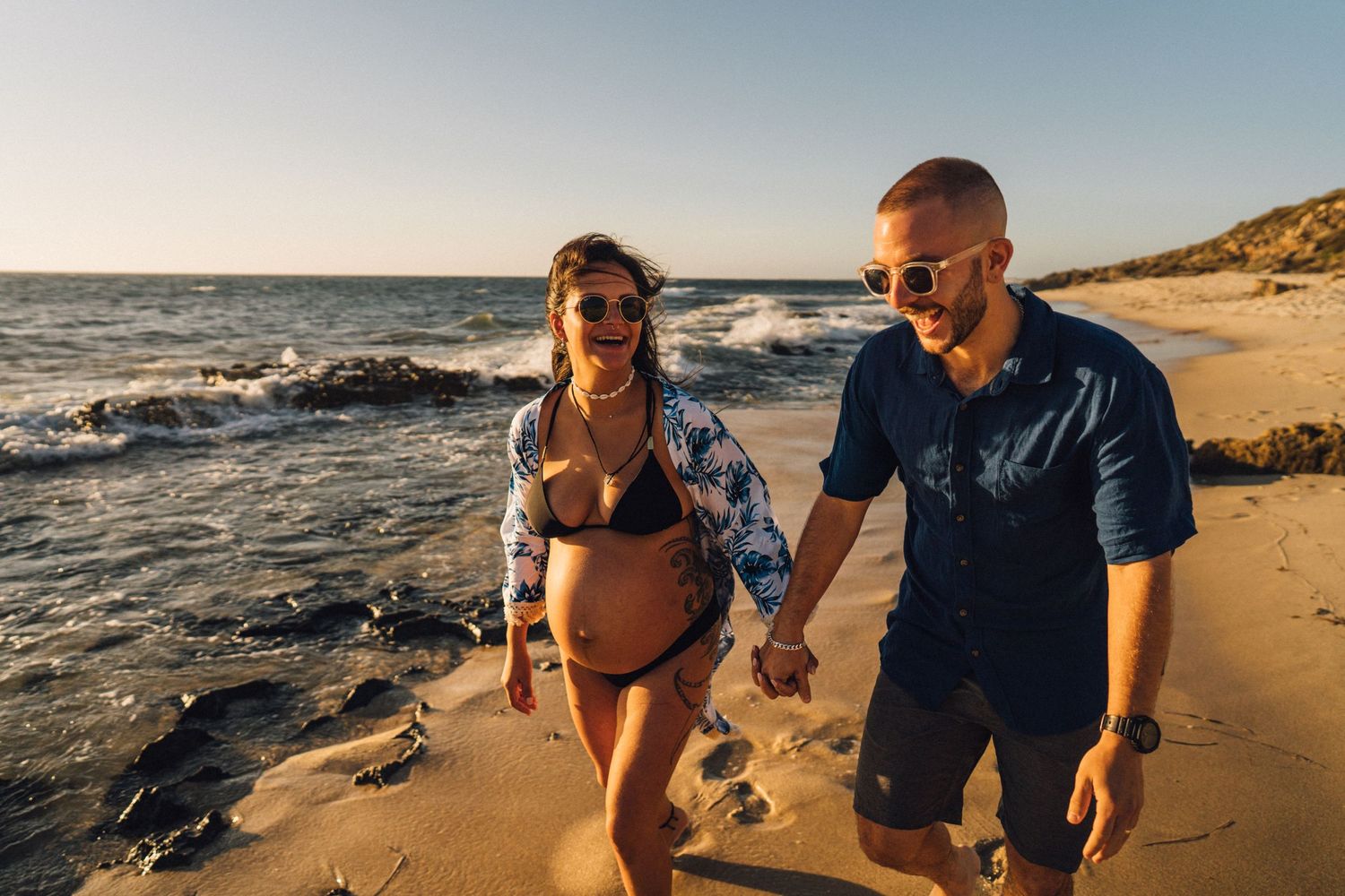A couple walks hand in hand along a rocky beach shore with waves crashing nearby.